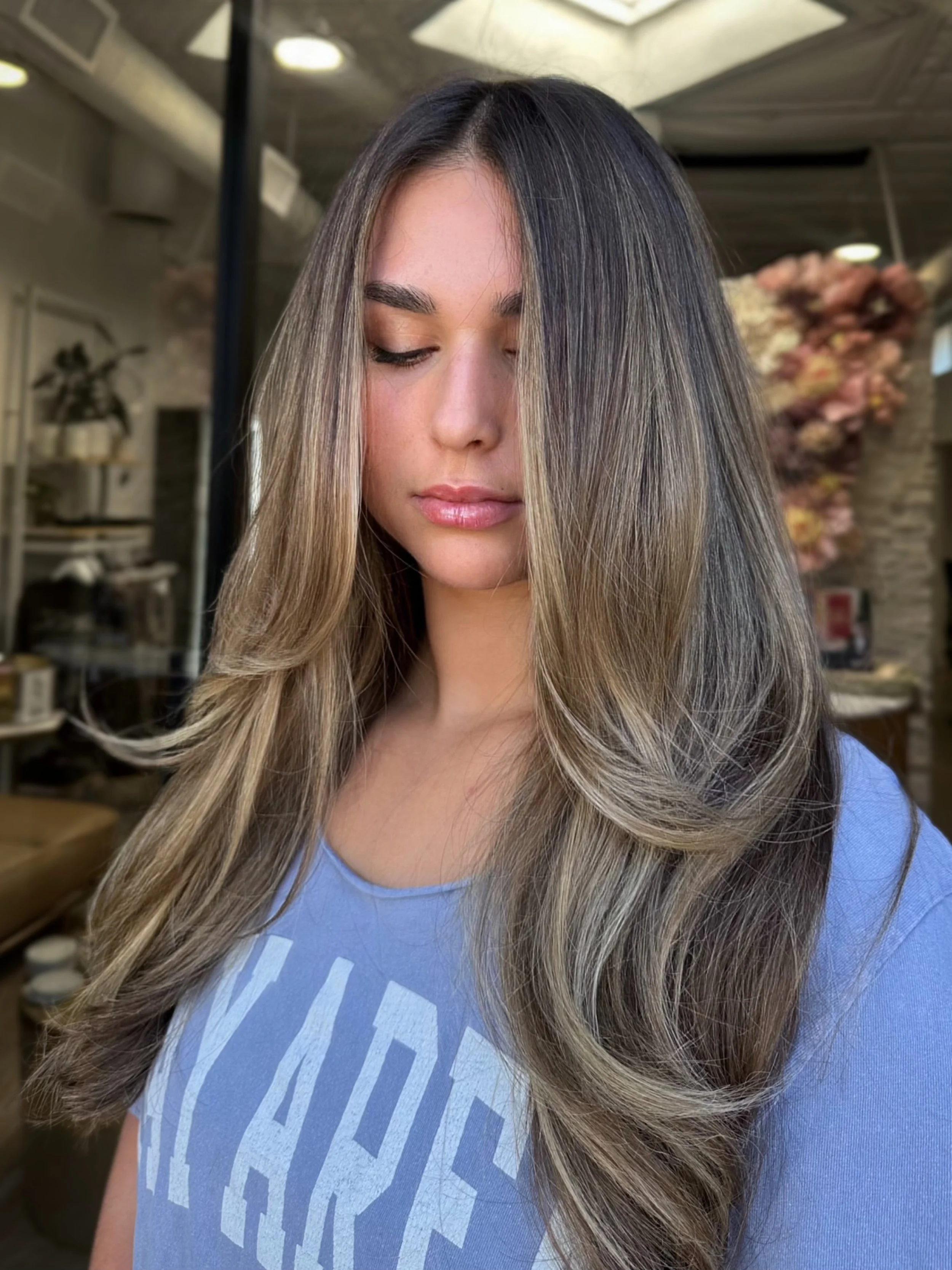 A woman with long, wavy blonde hair and light skin, with her eyes closed, standing inside a salon or studio with a background of shelves and decorative items.