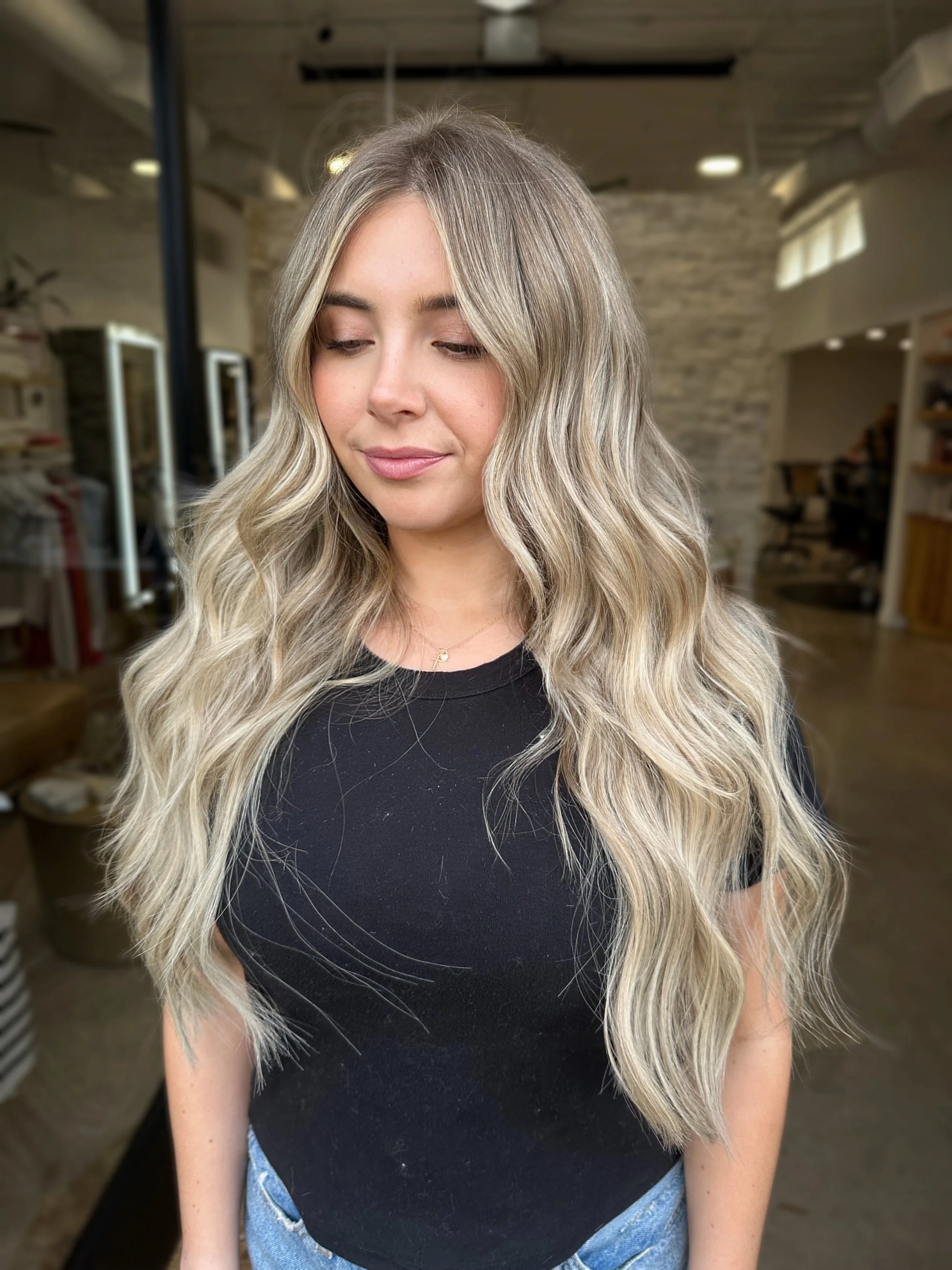 A woman with long, wavy blonde hair wearing a black t-shirt and blue jeans in a modern indoor space.