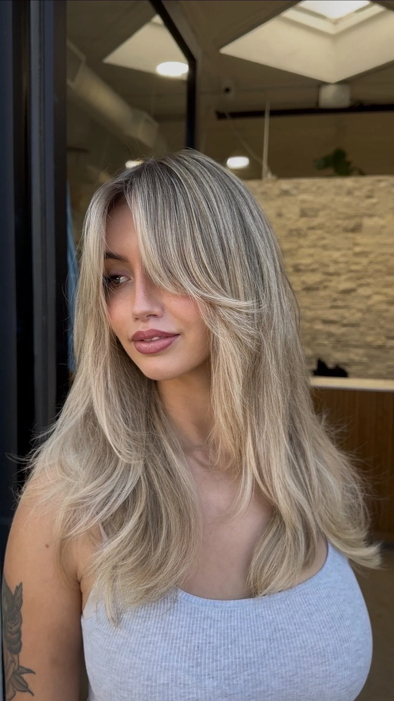 A woman with long blonde hair styled in loose waves, wearing a light blue tank top, standing indoors near a window or mirror, with a brick or stone wall and ceiling lights in the background.