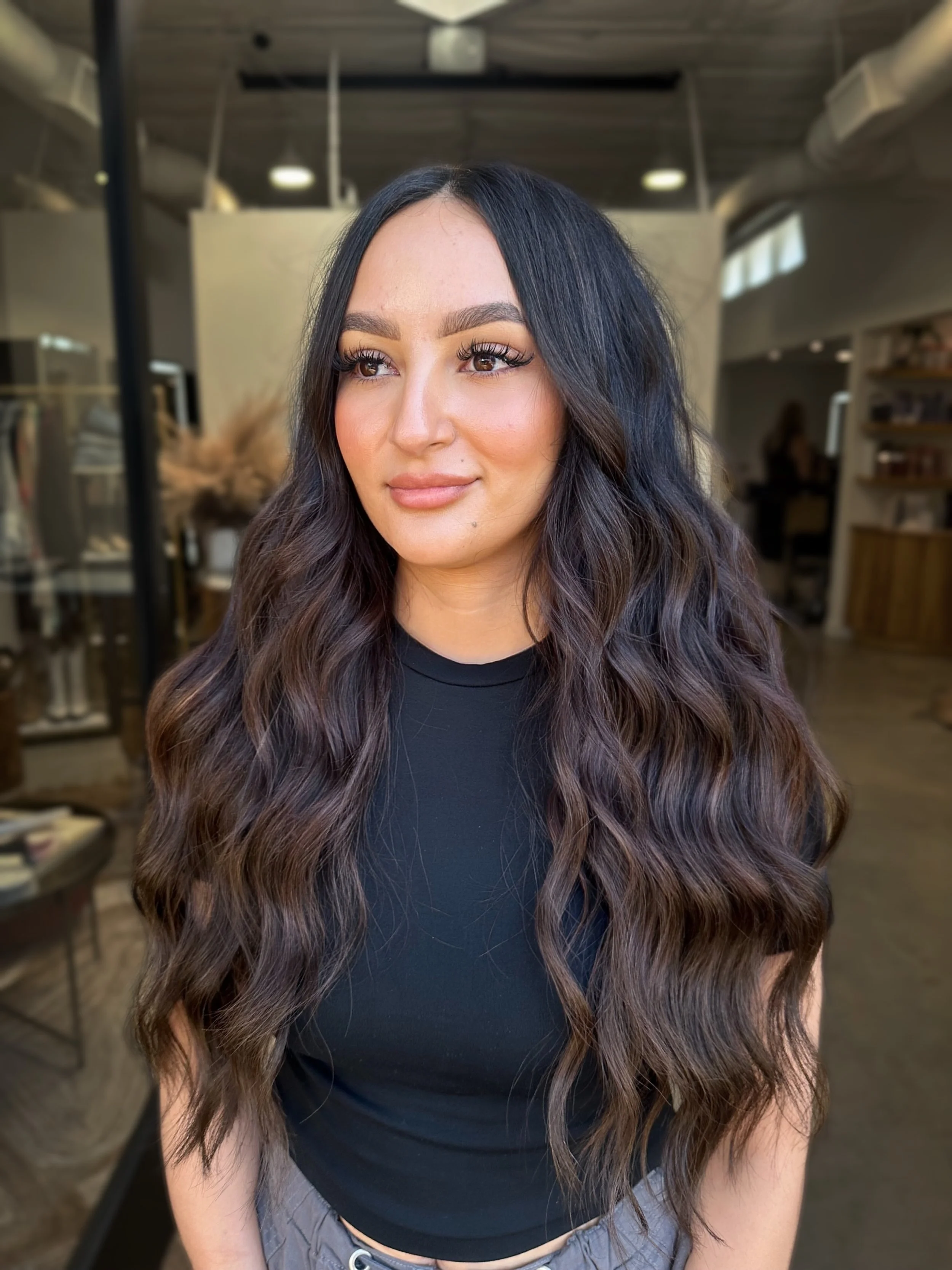 A woman with long, wavy dark hair and light skin wearing a black shirt and gray pants, standing in a well-lit indoor space with shelves and plants in the background.