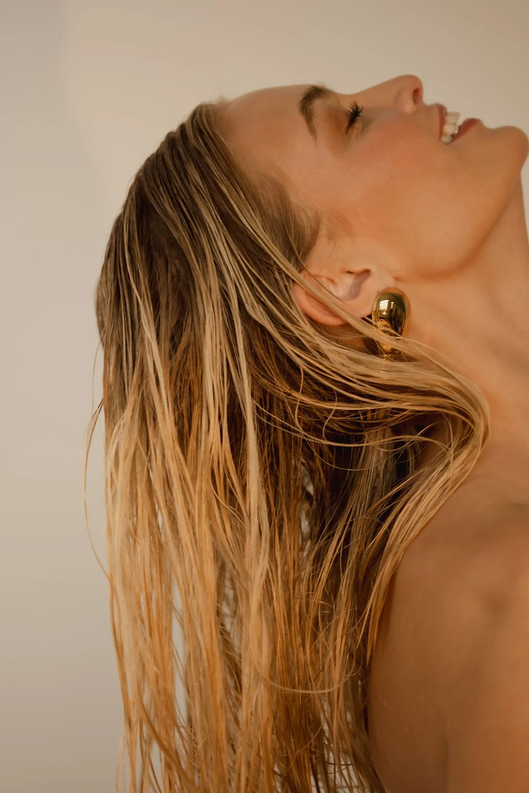 Close-up of a woman laughing, with wet blonde hair, wearing a large gold earring, against a plain light-colored background.