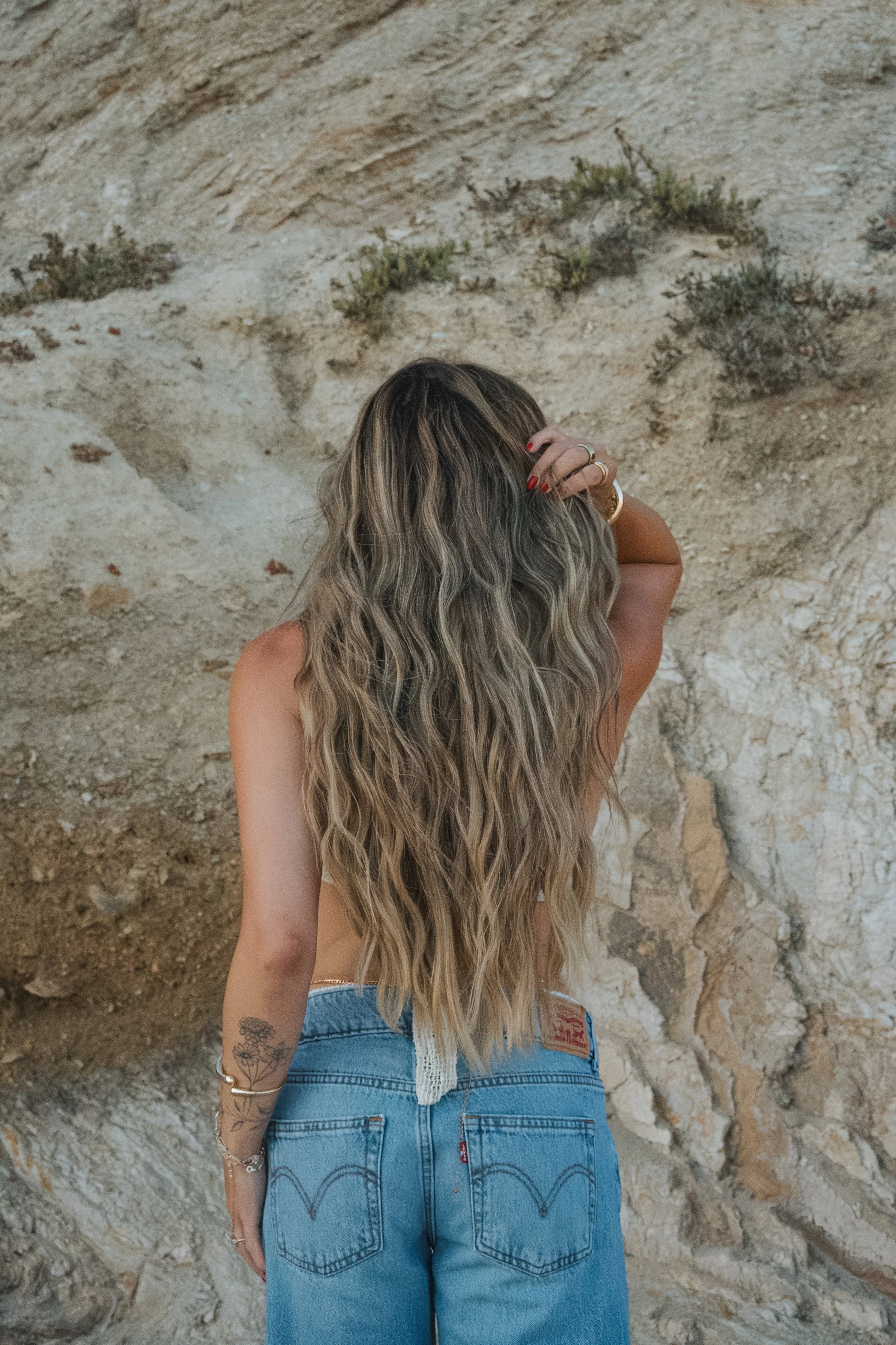 A woman with long wavy hair seen from behind standing in front of a rocky cliff, wearing blue jeans and a bracelet on her left wrist.