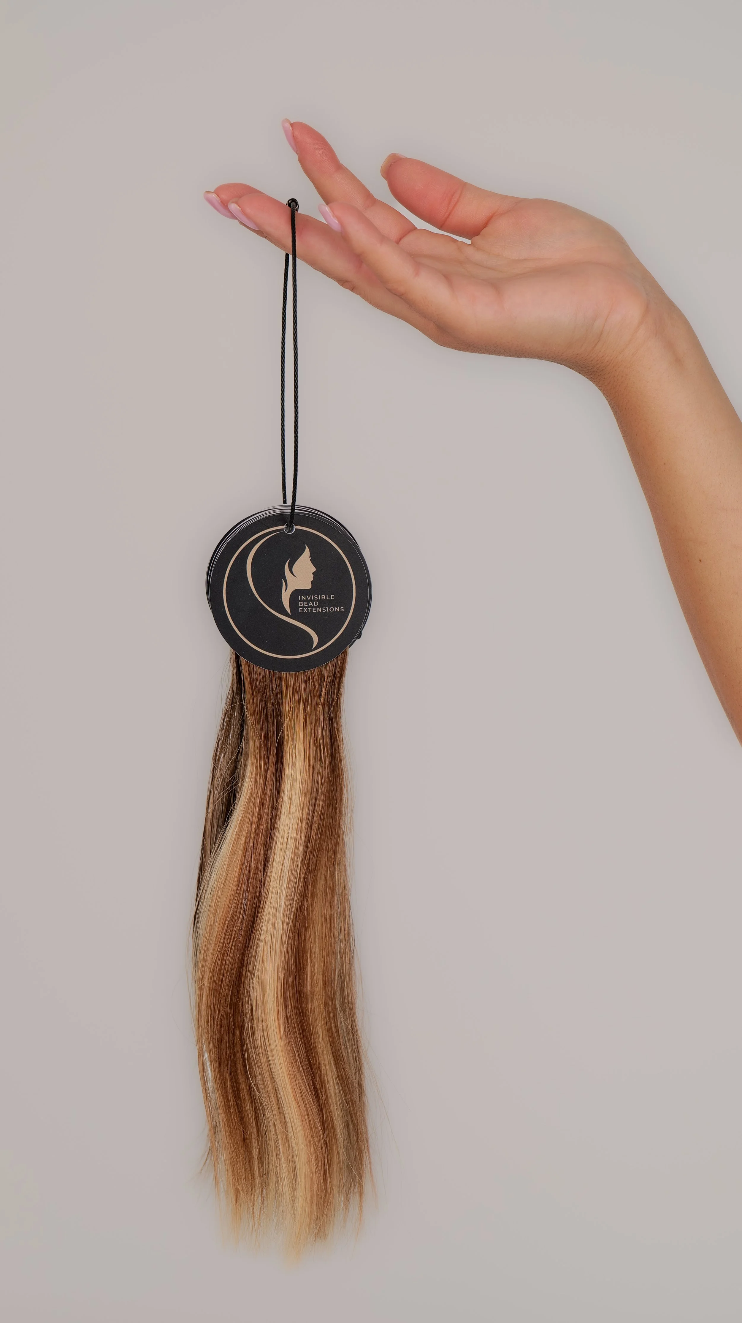 Hand holding a black tag with a silhouette of a face and the text 'Invisible Bead Extensions' attached to a long strand of straight, ombré hair.