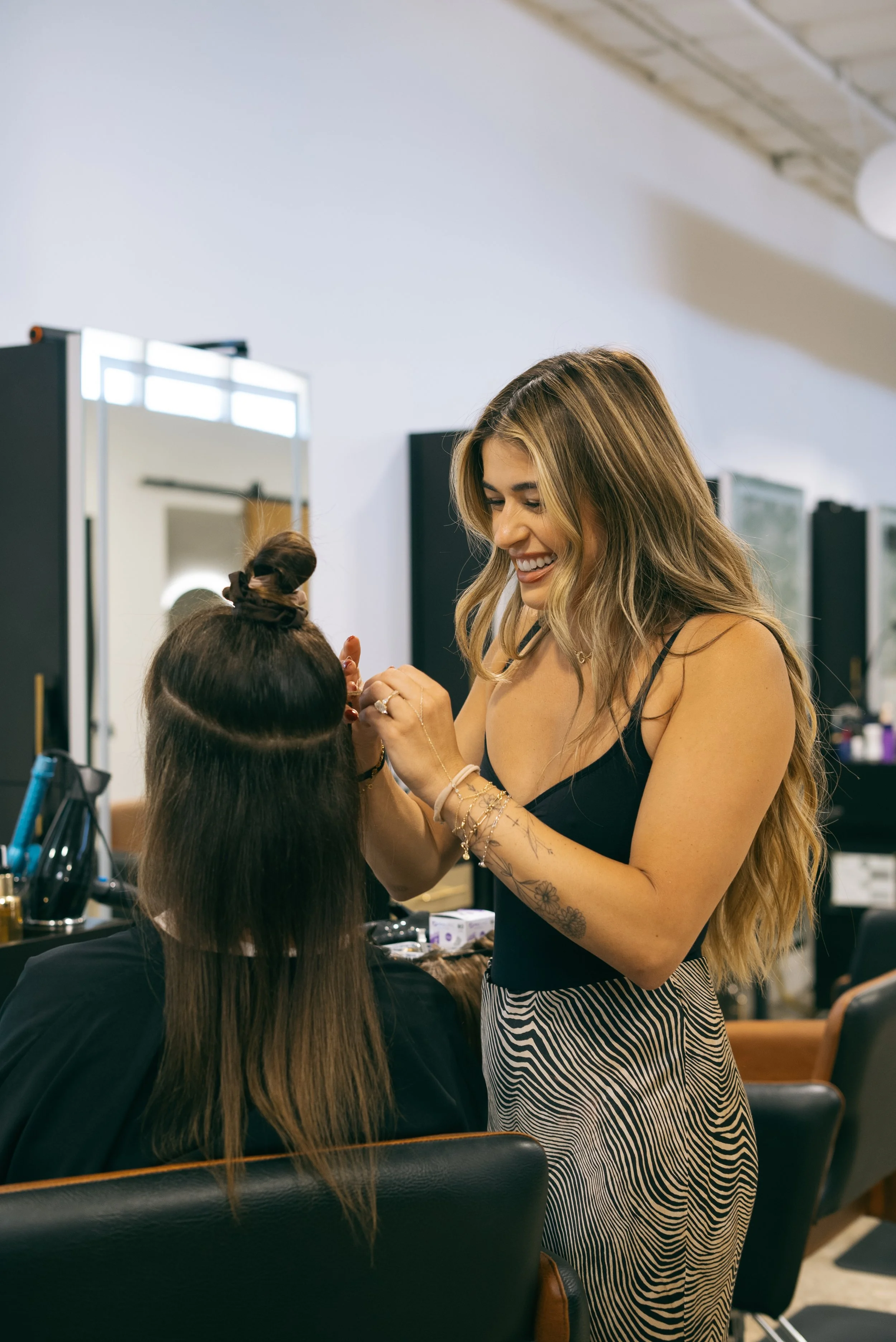 Woman with long blonde hair applying makeup to a seated client in a salon.