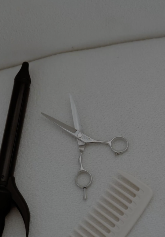 A pair of small, silver scissors, a white plastic comb, a black hairdryer, and a white wall with a curved ceiling.