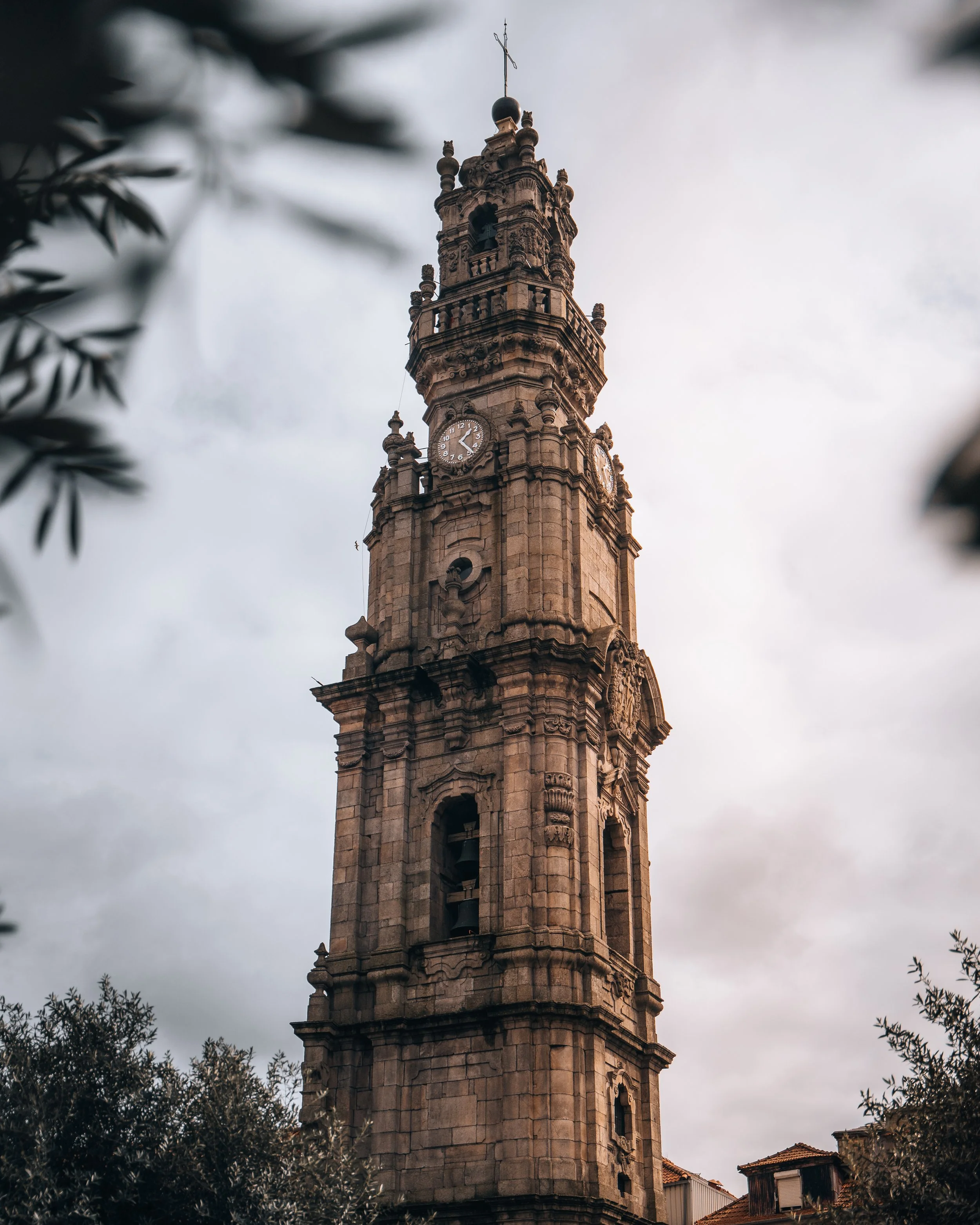 A historic clock tower with a detailed stone facade, topped with a cross, against a cloudy sky with some tree branches in the foreground.