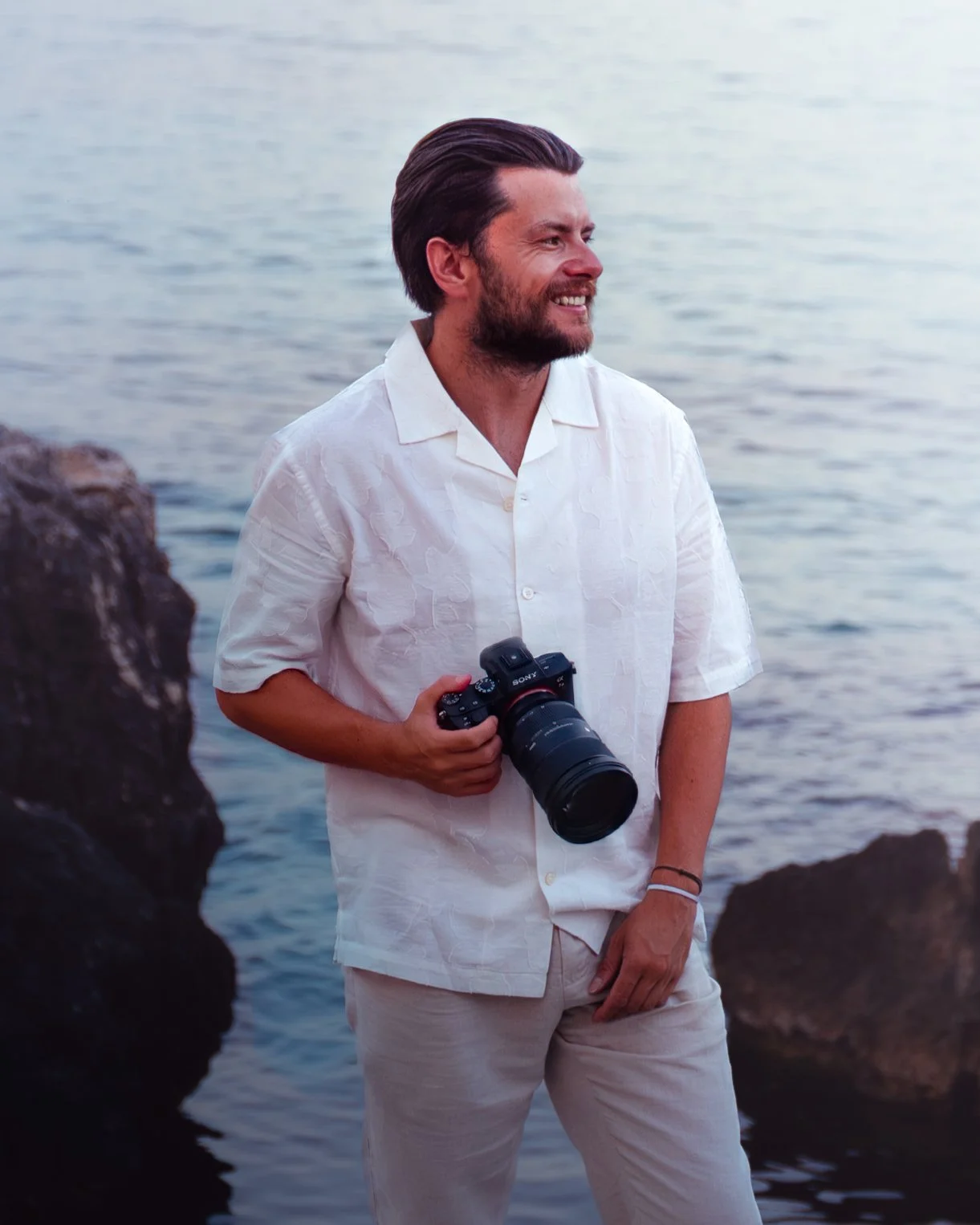 A man standing by the water holding a camera, wearing a white shirt and beige pants, smiling and looking to the side, with rocks and water in the background.