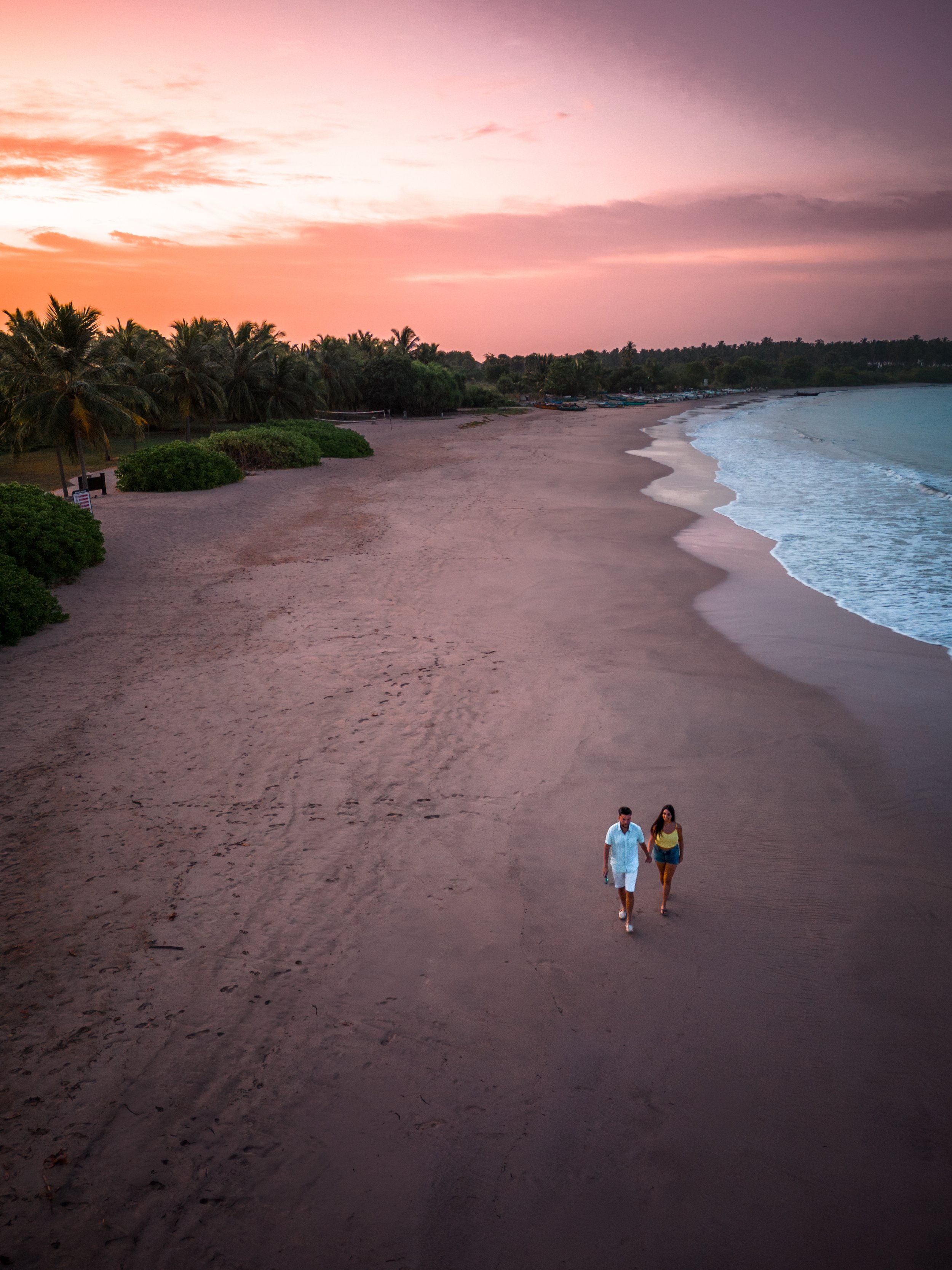 A couple walking along a sandy beach during sunset with palm trees and boats in the background.