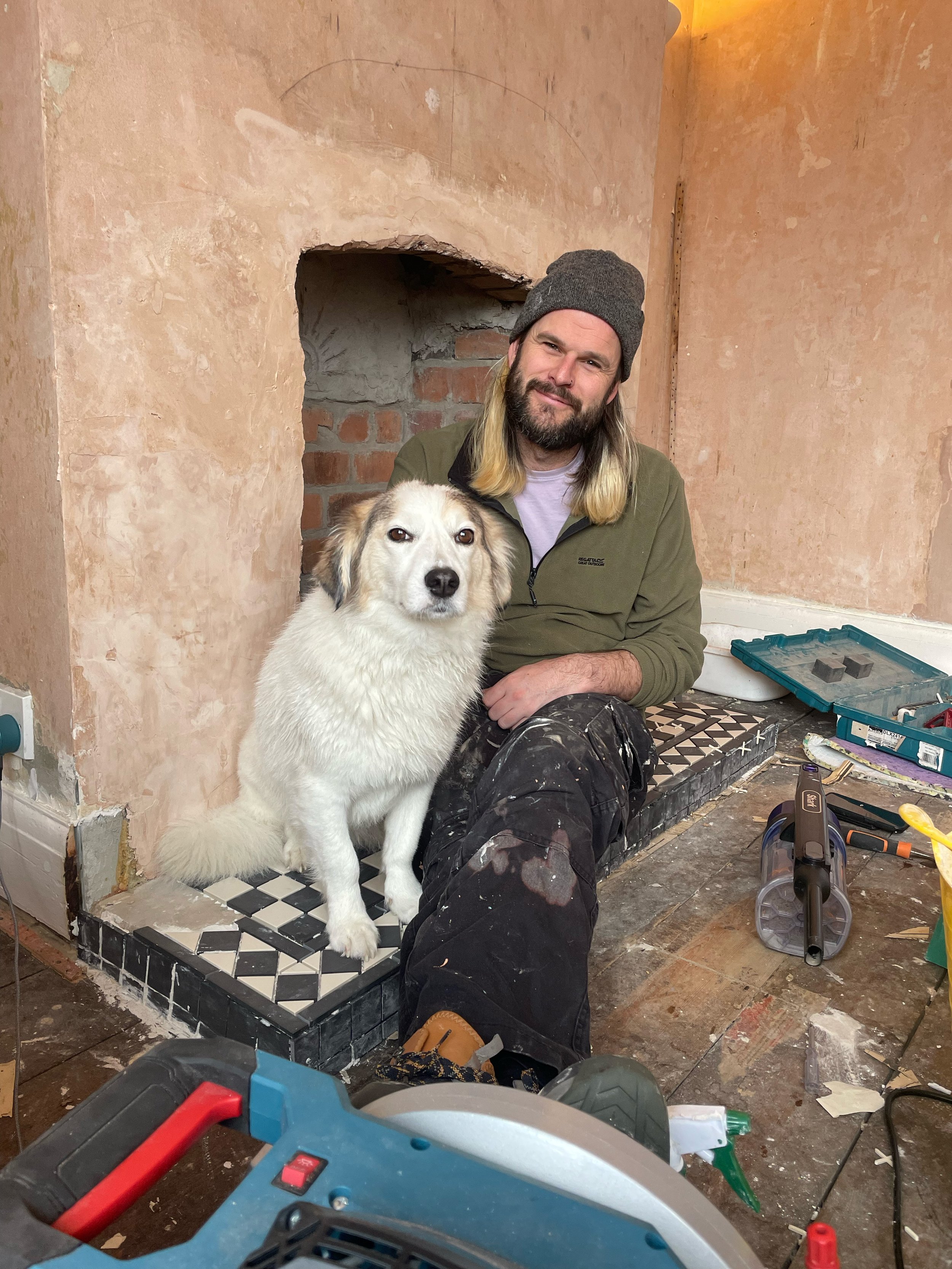 A man with long hair and a beard wearing a gray beanie and green jacket is sitting next to a white dog with brown markings, inside a room under renovation with construction tools and materials around. The man is smiling and there is a partially finished fireplace or wall opening behind them.