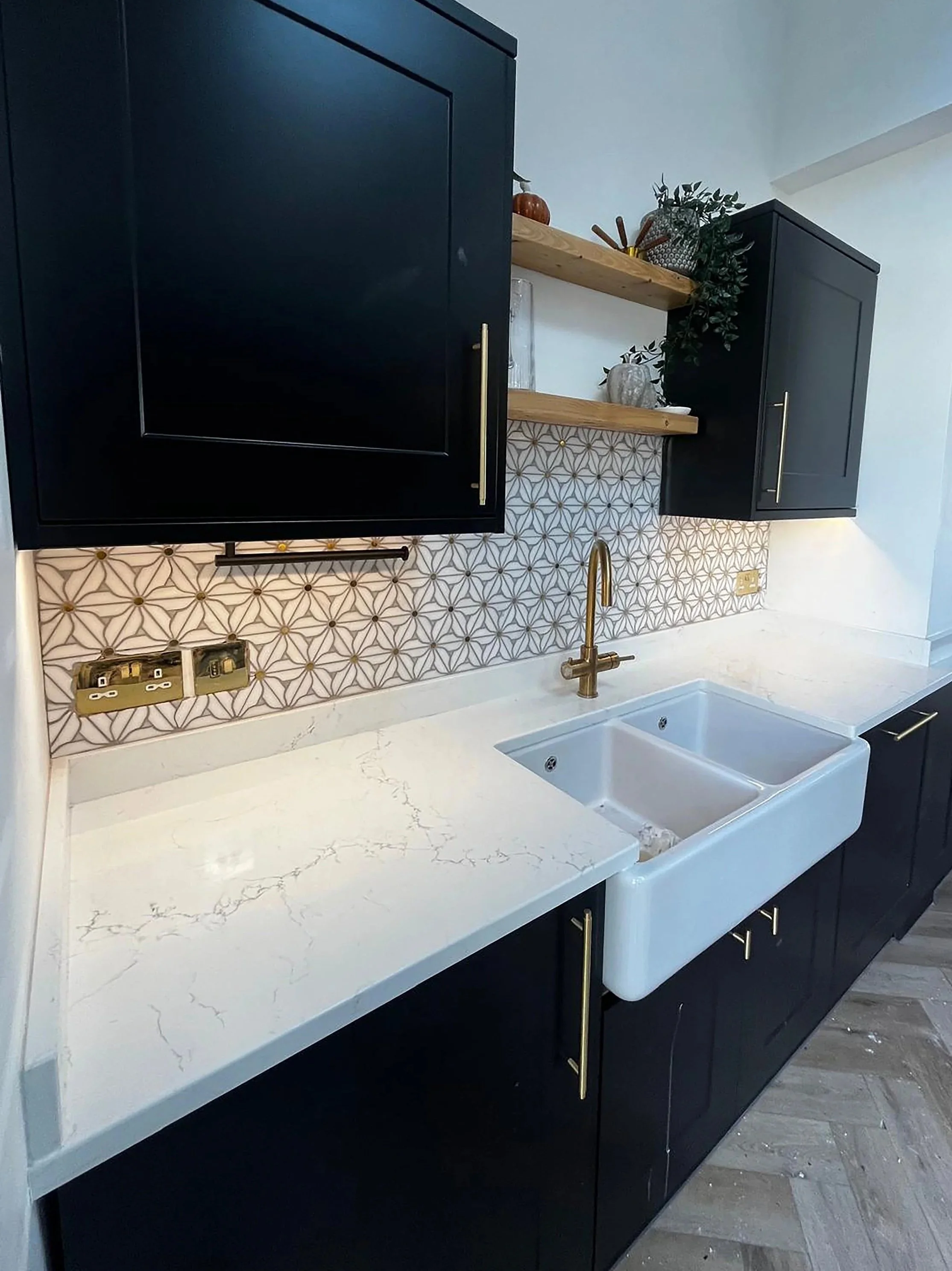 A modern kitchen featuring black cabinetry with brass handles, a white marble countertop with subtle veining, a white farmhouse sink, a gold faucet, and open wooden shelves decorated with plants, vases, and small decorative items.