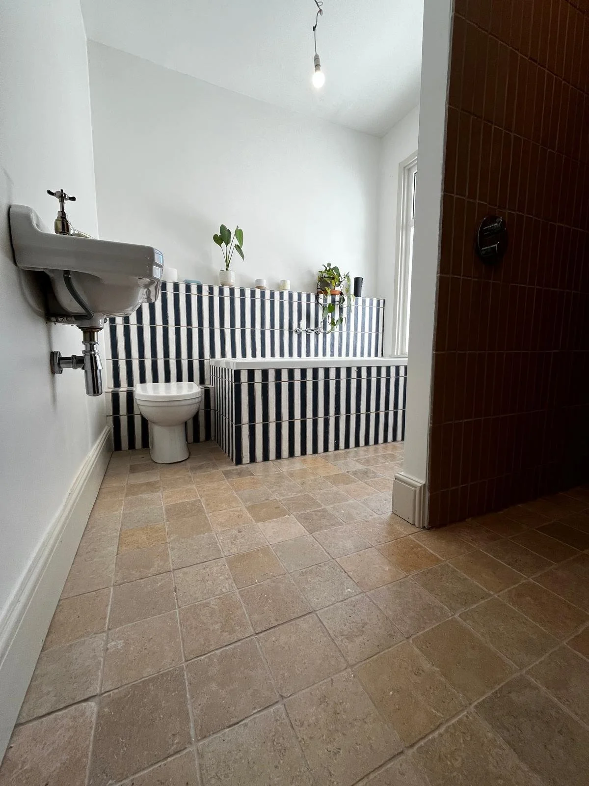 Bathroom with a striped black and white bathtub, a toilet, small plants on the tub, and a white sink. Brown tiled wall partially visible to the right, beige tiled floor, white walls, and a ceiling light.