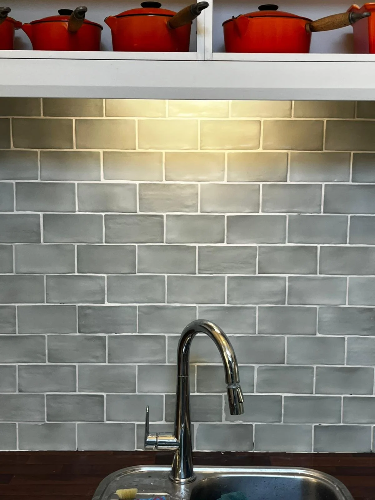 Red pots with wooden-handled ladles on a white shelf above a kitchen sink with a chrome faucet, gray tiled backsplash, and dark wooden countertop.