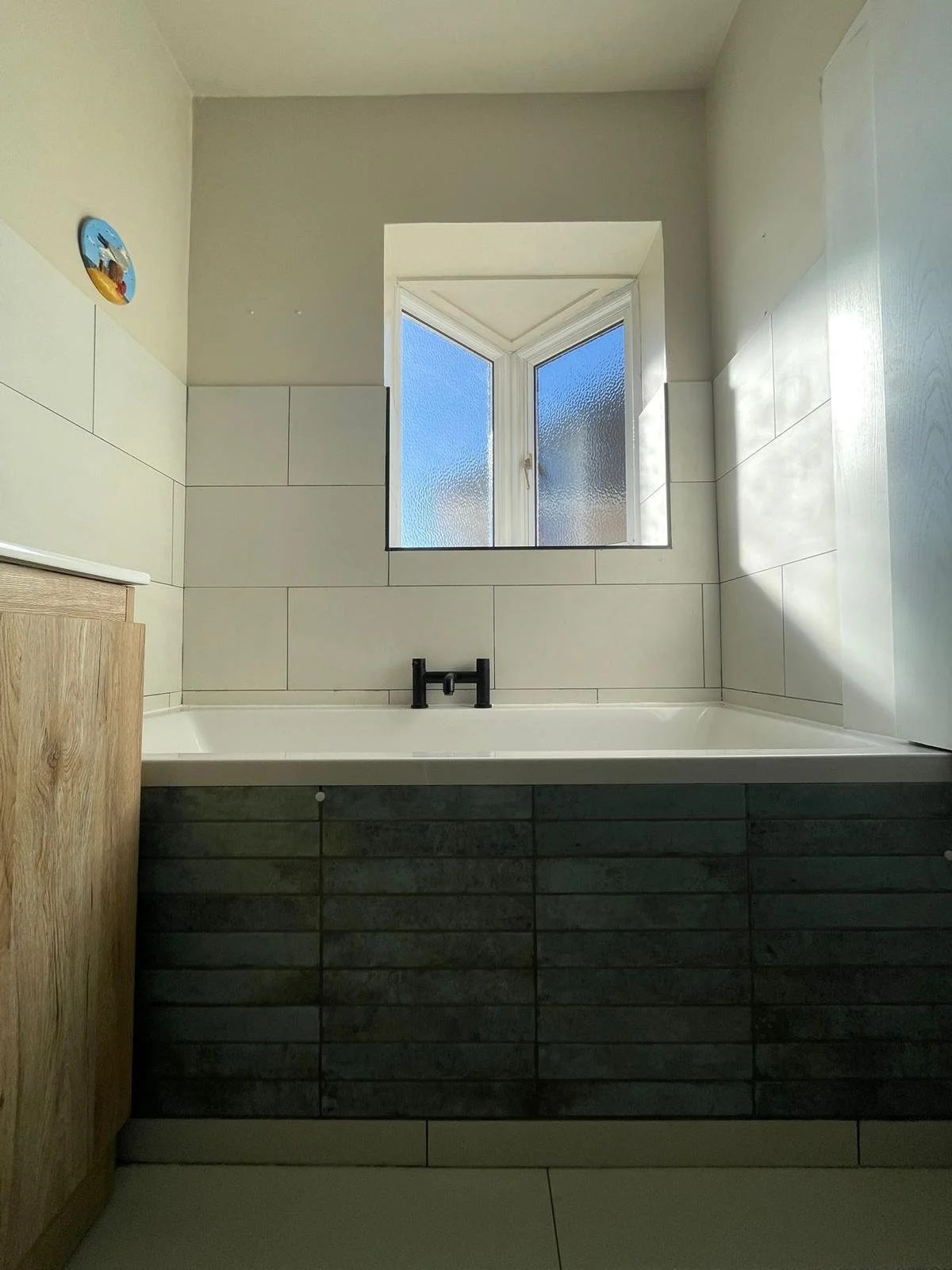 Bathroom with a bathtub under a window, black faucet, tiled walls, and a wooden cabinet on the left.