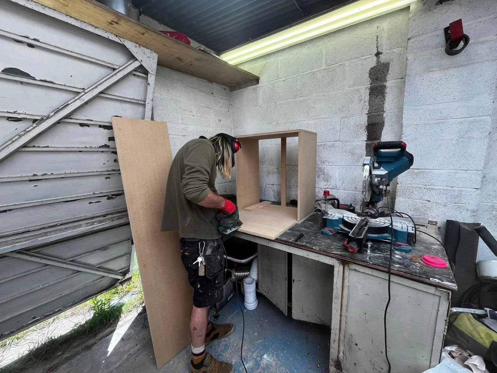 A person working in a workshop, using a power tool on a wooden structure placed on a workbench. The workshop has concrete walls, a metal roof, and various tools and supplies around.