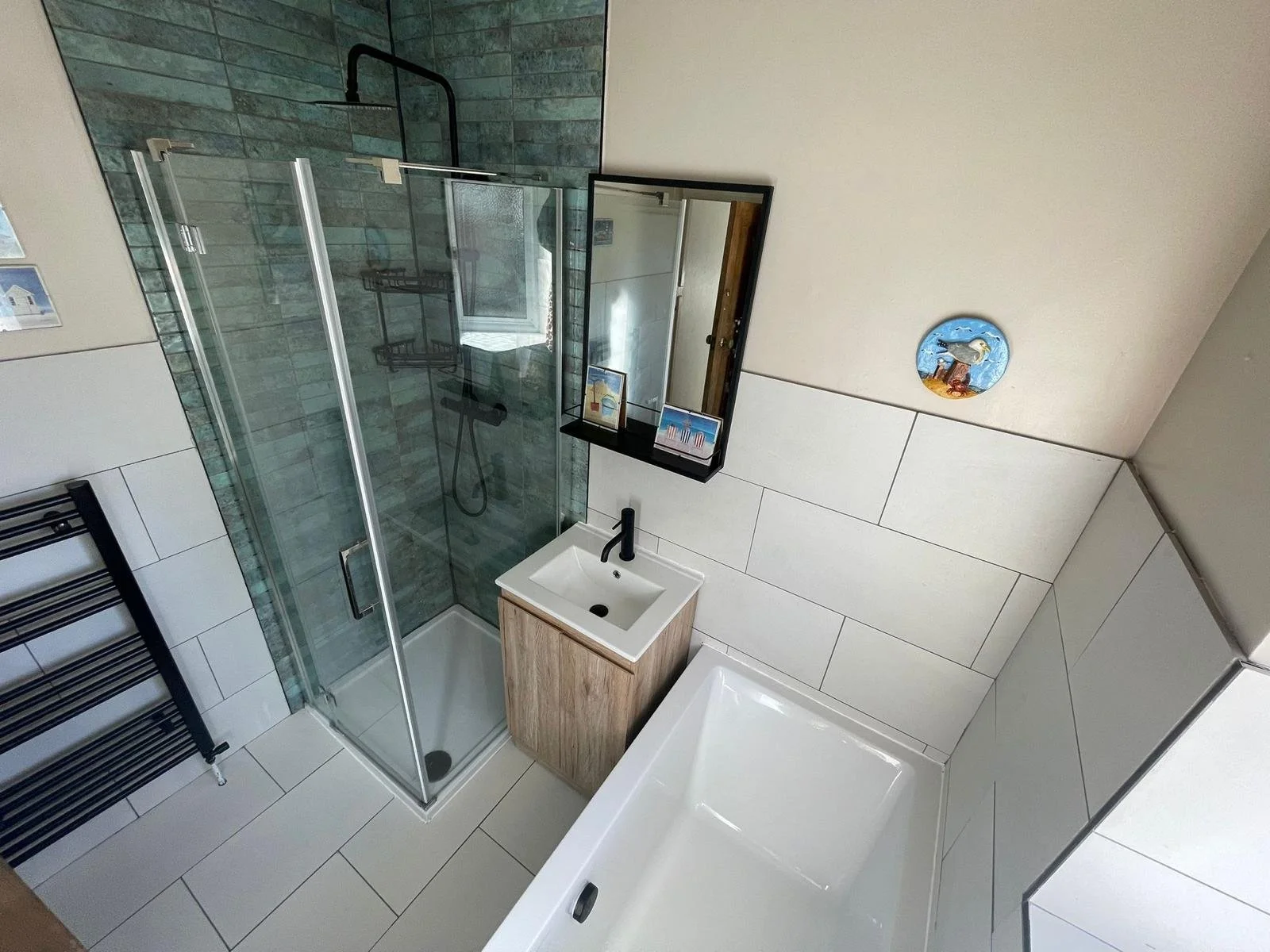 Modern bathroom with glass-enclosed shower, white bathtub, small wooden vanity with white sink, black faucet, large mirror, black towel radiator, decorative plate on the wall, and framed artwork.