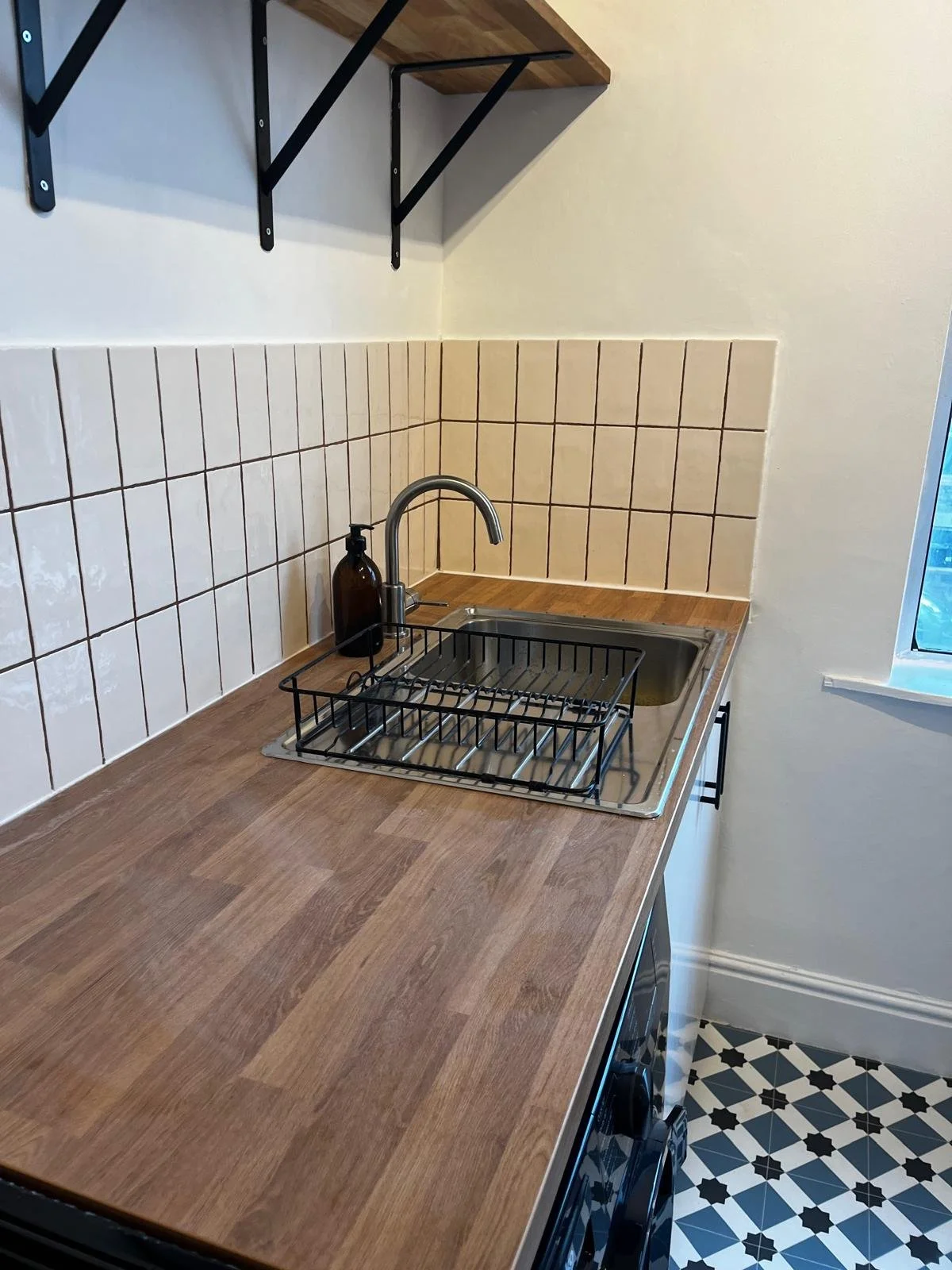 Small kitchen corner with wooden countertop, stainless steel sink, black metal dish rack, brown soap dispenser, beige tiled backsplash, white wall, small floating wooden shelf, black metal brackets, and a window with a black frame. Floor has black and white patterned tiles.