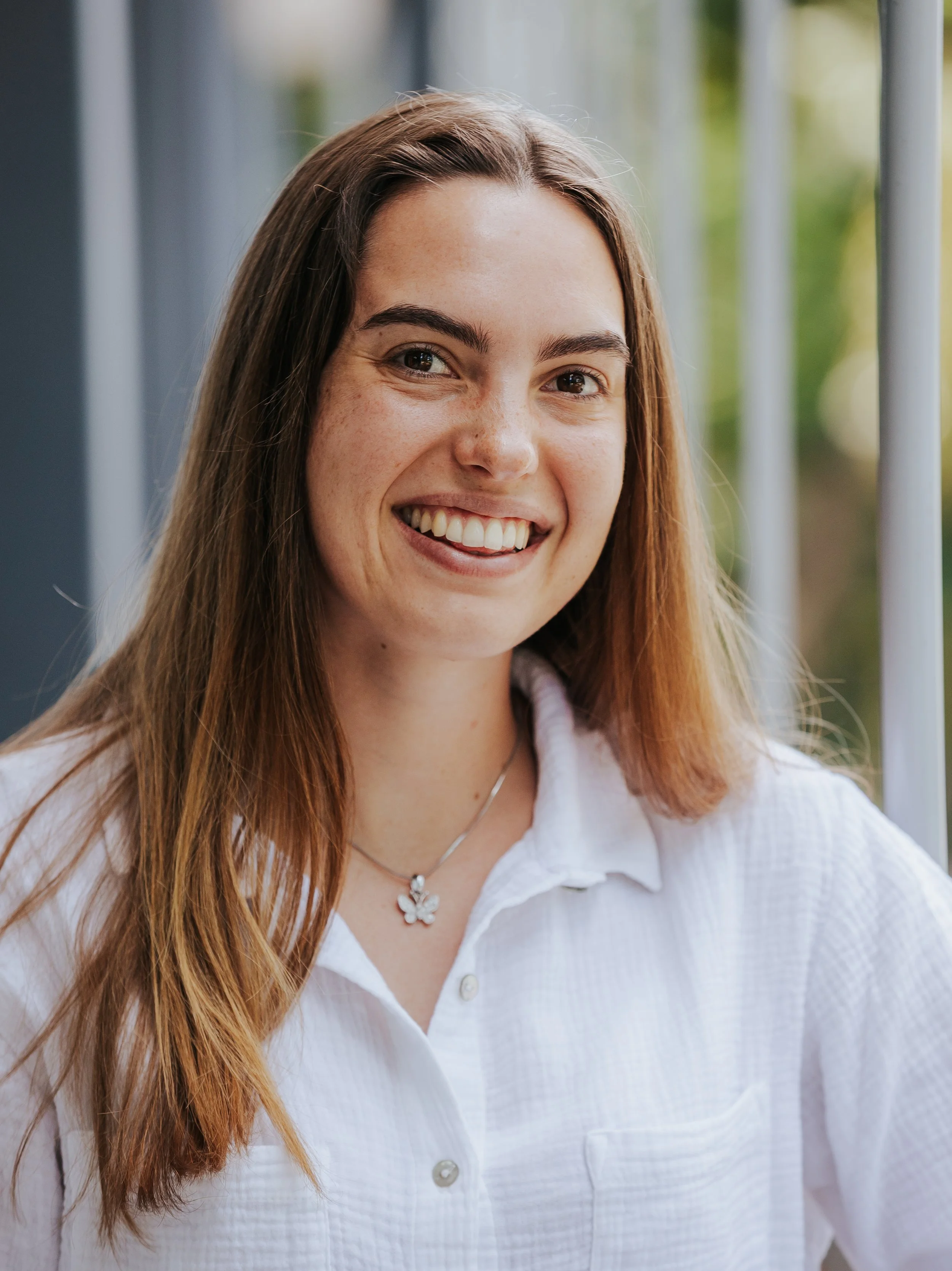 A smiling young woman with long, light brown hair, wearing a white collared shirt, and a butterfly-shaped necklace, standing outdoors with visible green blurred foliage in the background.