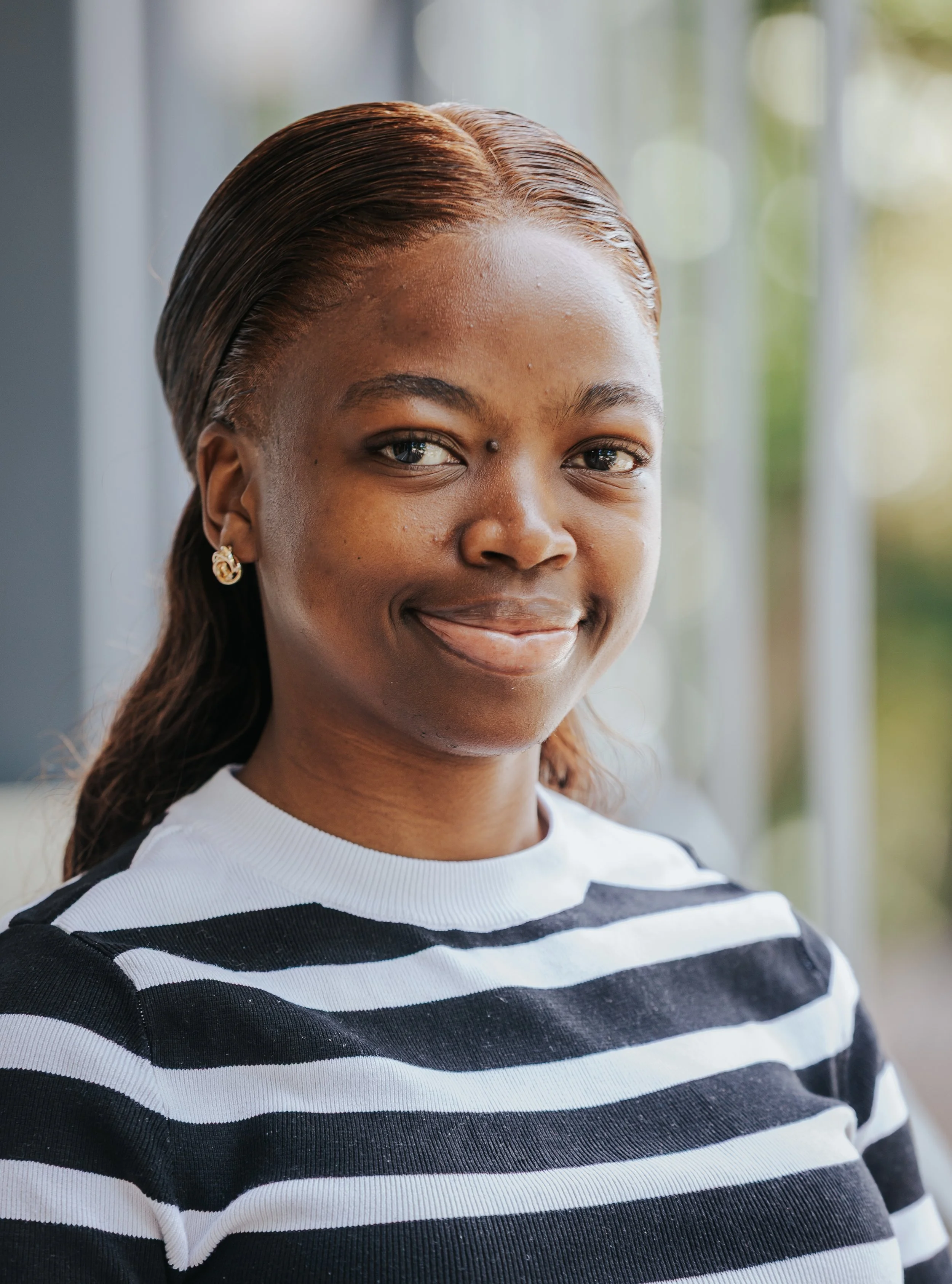 A woman with brown hair styled with a side part, smiling slightly, wearing a black and white striped shirt, gold earrings, and standing near a window with a blurred outdoor background.