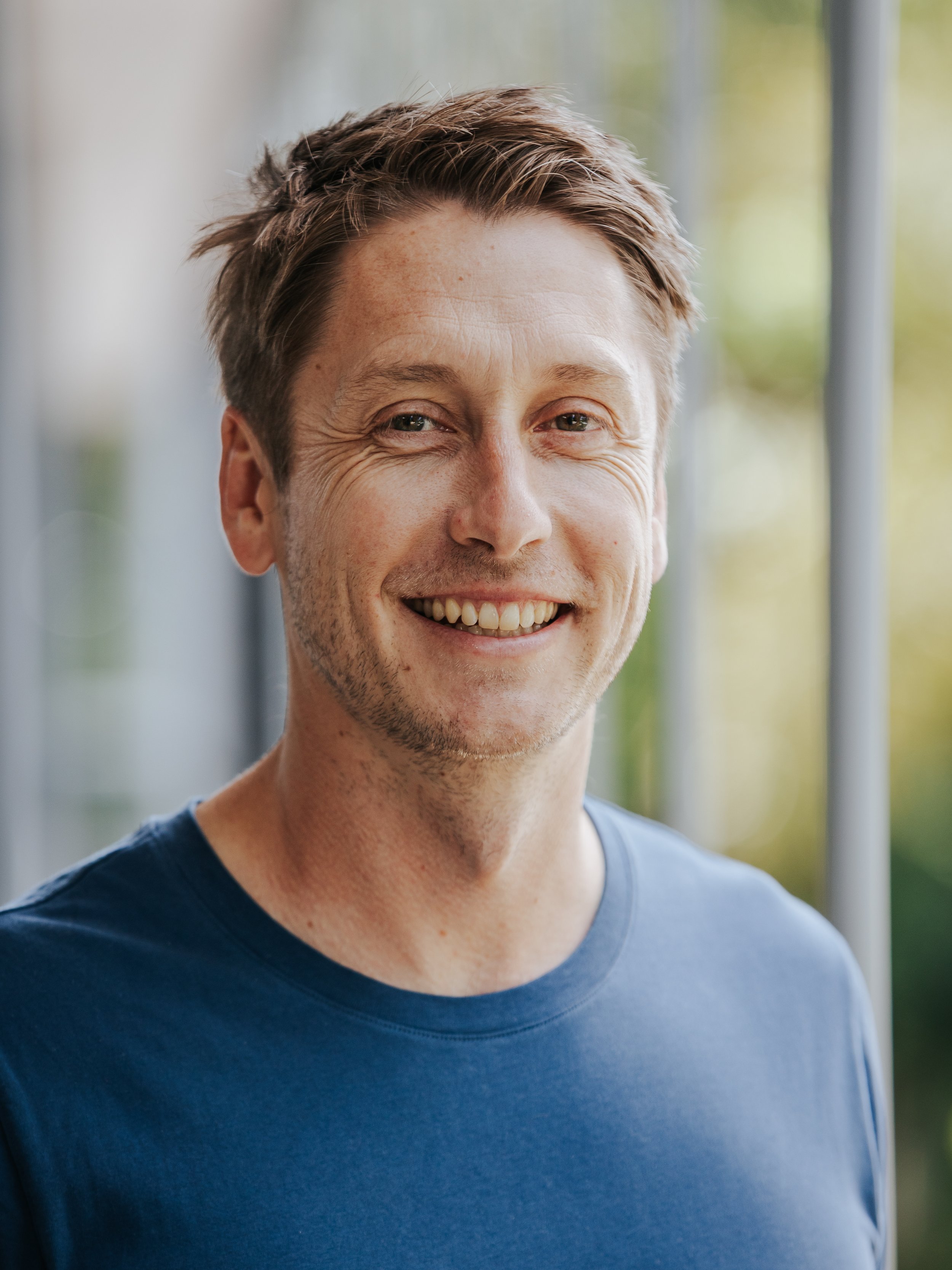 Smiling man with short brown hair wearing a blue T-shirt, standing indoors near a window with blurred greenery background.