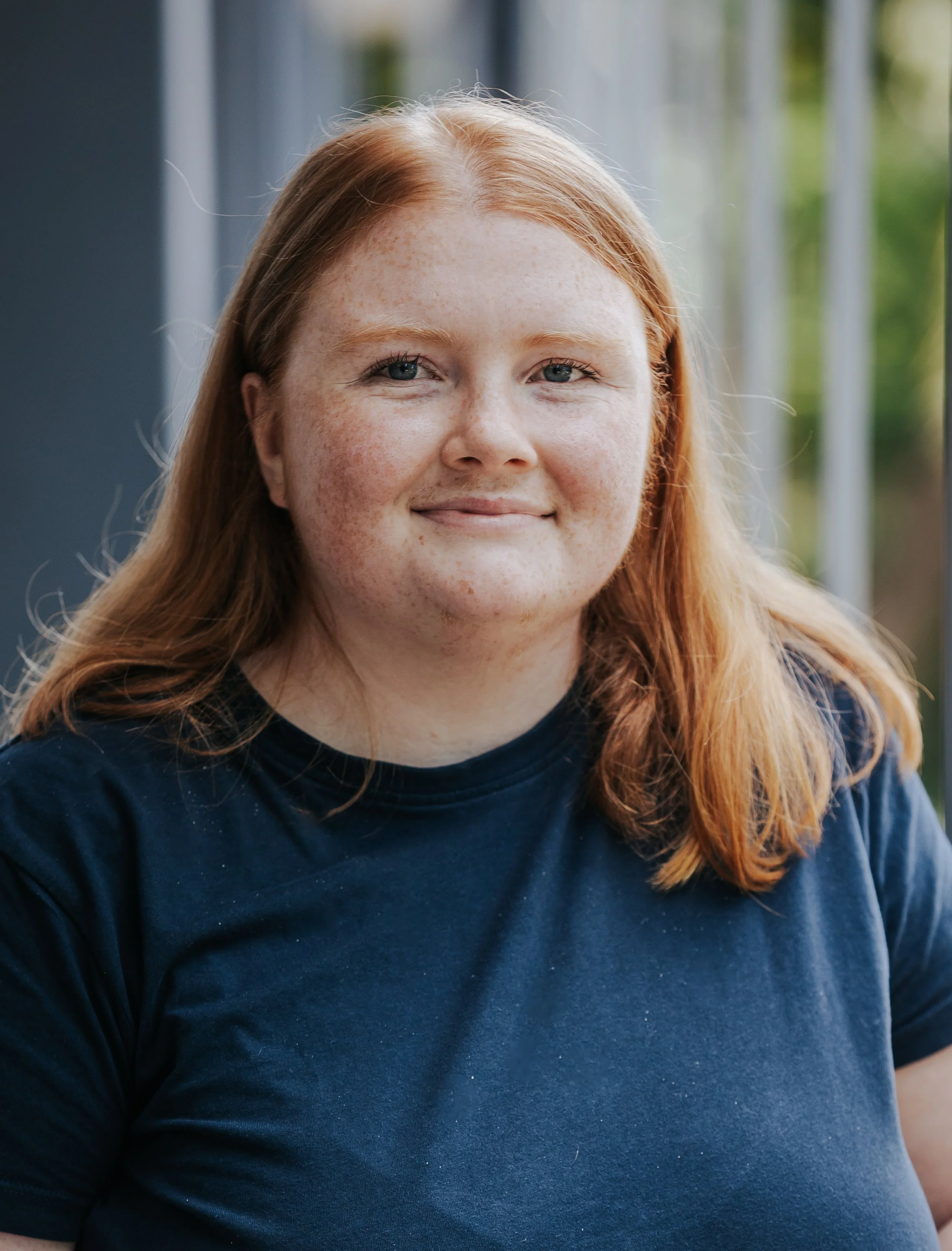 Close-up of a woman with red hair, blue eyes, freckles, smiling, wearing a dark blue shirt, outdoors with blurred green and white background.