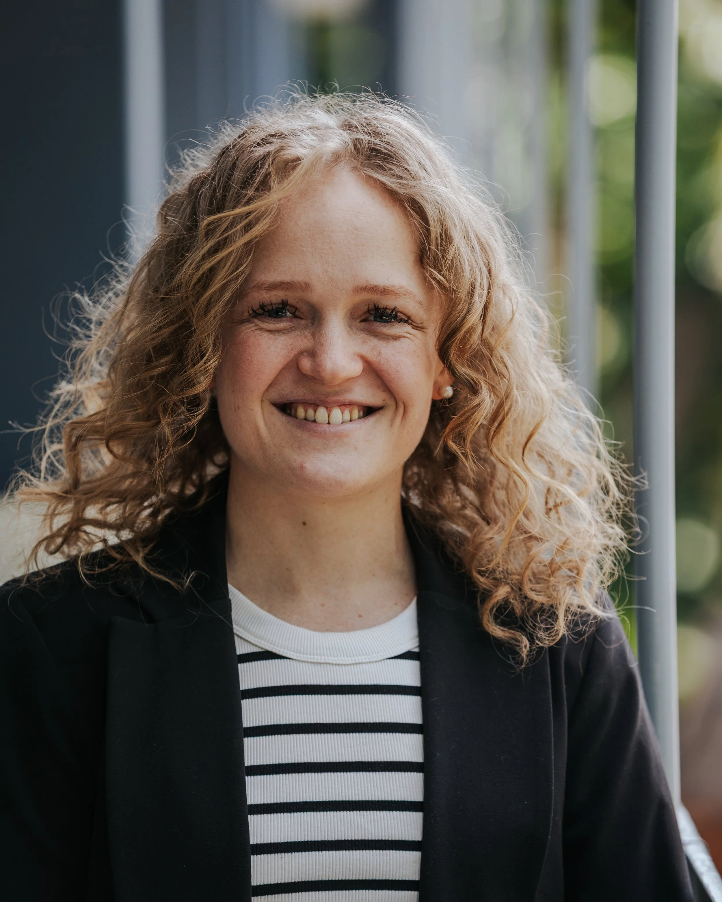 Close-up of a woman with curly blonde hair, smiling, wearing pearl earrings, a black jacket, and a striped shirt, outdoors with blurred green background.