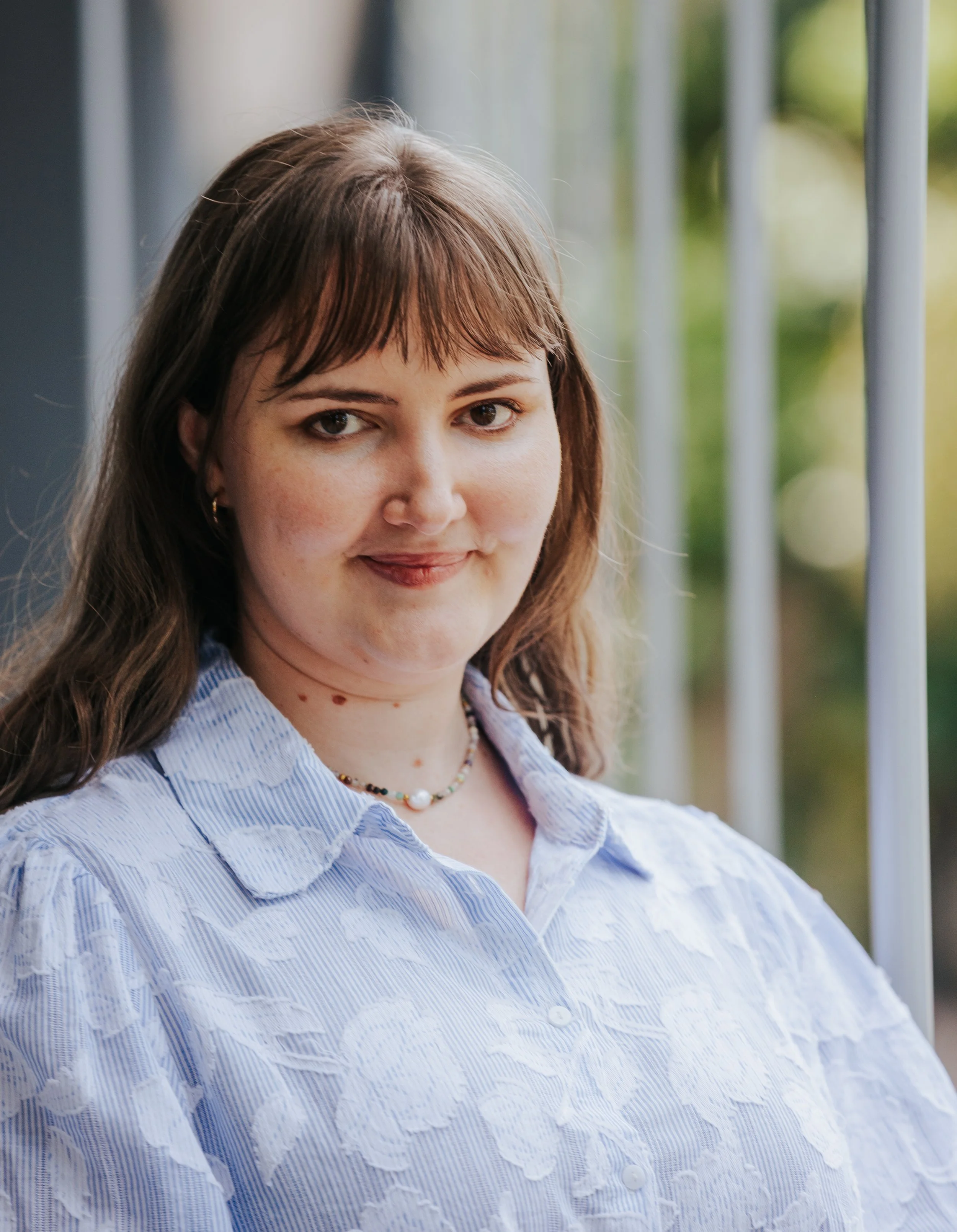 A young woman with brown hair and bangs wears a light blue and white striped shirt and a colorful beaded necklace. She is outdoors near a metal railing, with blurred green foliage in the background.