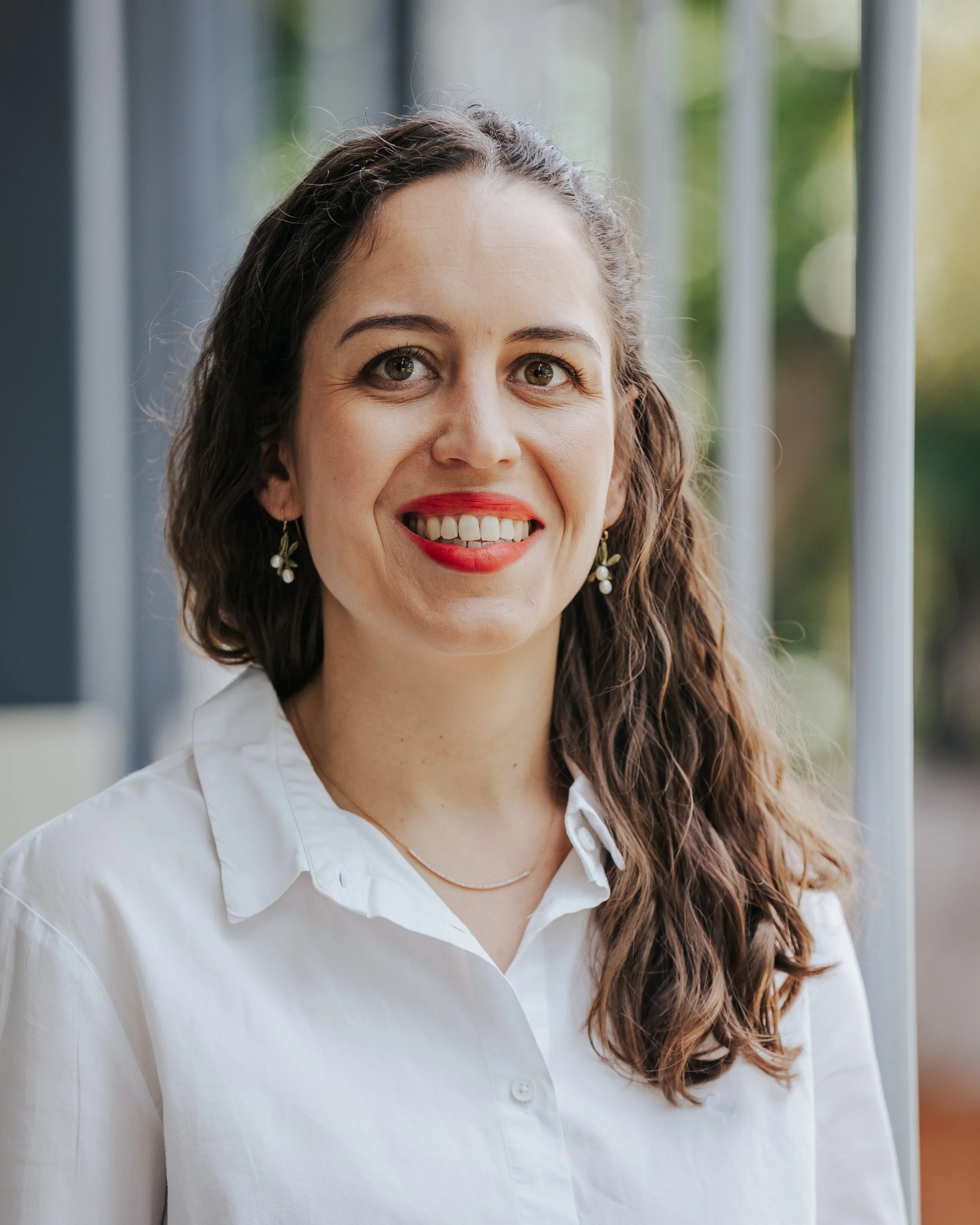 A woman with long, curly brown hair, green eyes, and light skin. She is smiling, showing her teeth, and wearing red lipstick, earrings with flowers, a delicate gold necklace, and a white collared shirt. The background is blurred greenery and vertical metal bars.