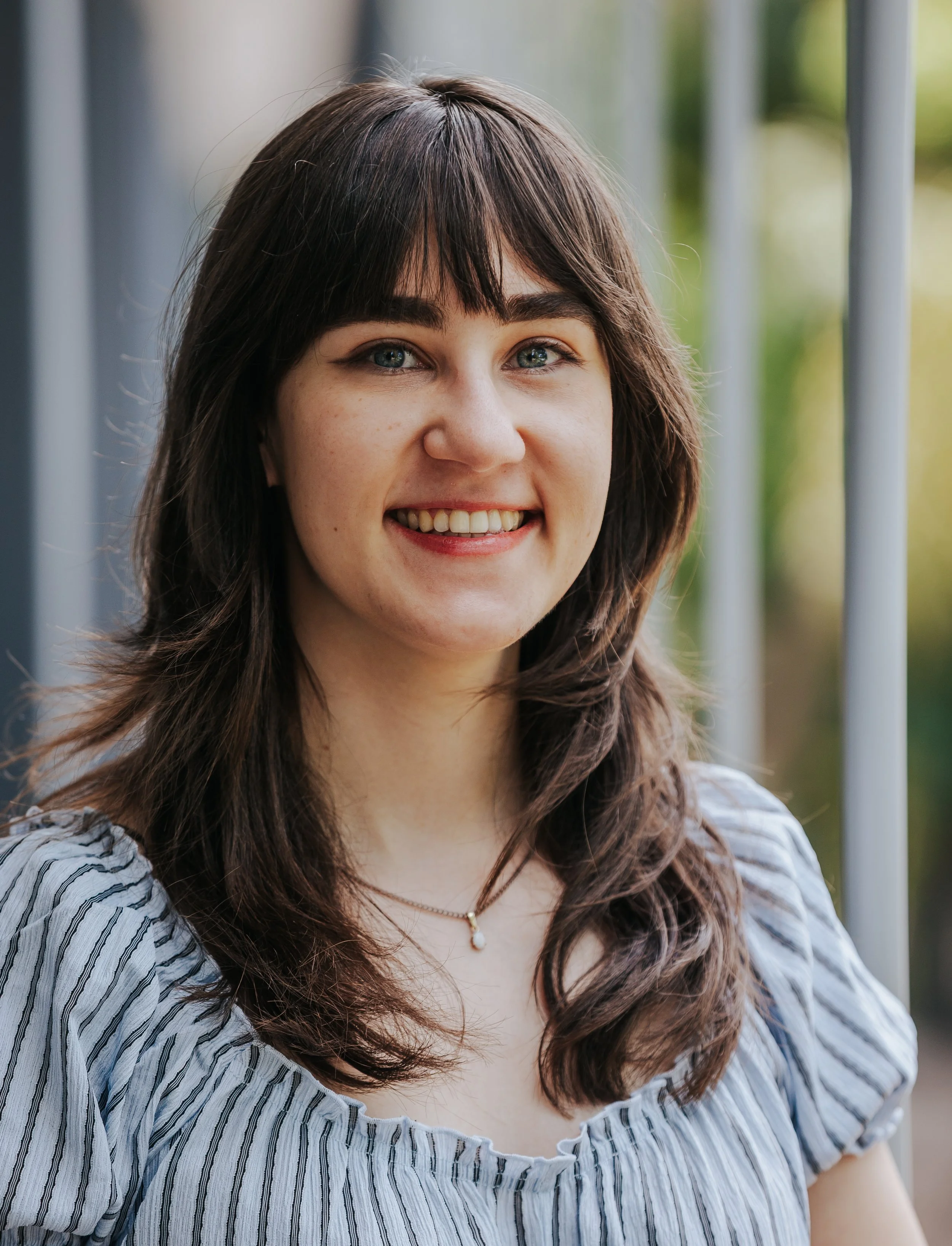 Close-up of a smiling woman with long wavy brown hair, wearing a striped blue and white blouse, standing outdoors near a metal railing, with blurred green foliage in the background.