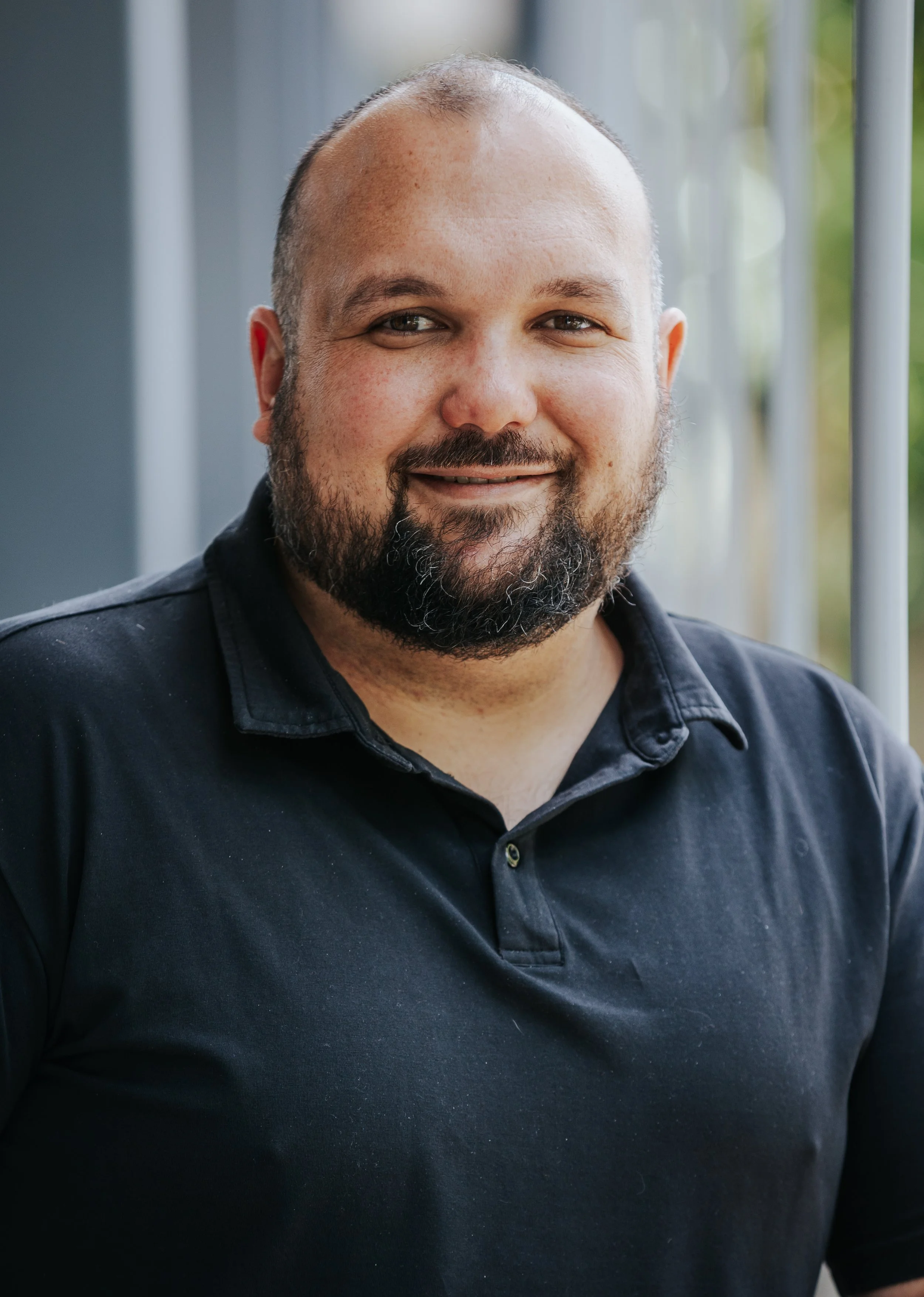 A smiling man with a beard and mustache, wearing a black collared shirt, standing outdoors near a metal fence with blurred greenery in the background.
