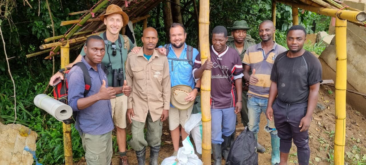 Group of nine men standing together outdoors in front of a bamboo structure, smiling at the camera. One man is giving a thumbs-up, and another has binoculars around his neck. They are dressed in casual clothing and outdoor gear.