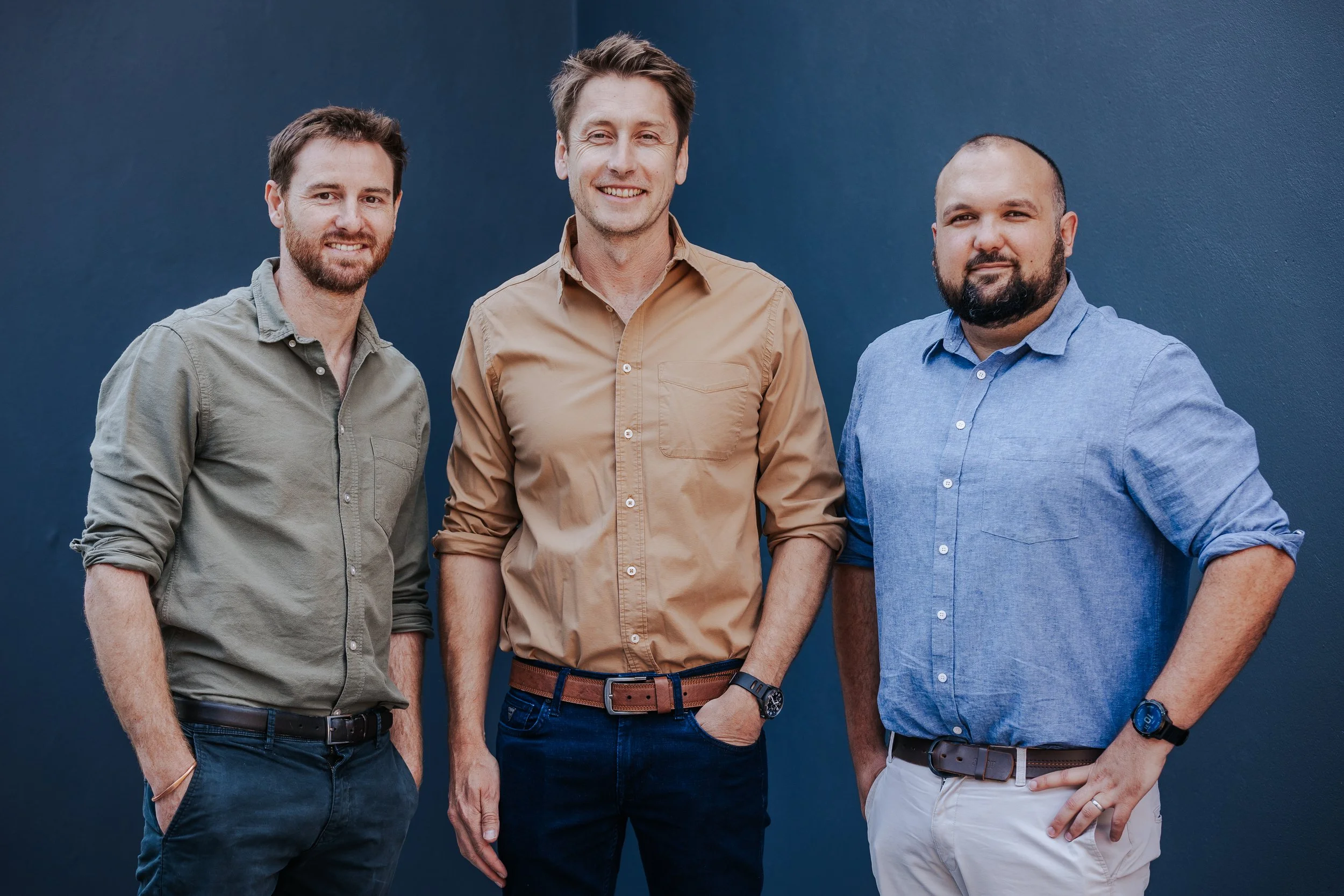 Three men standing against a dark blue wall, smiling and looking at the camera, dressed in casual shirts.