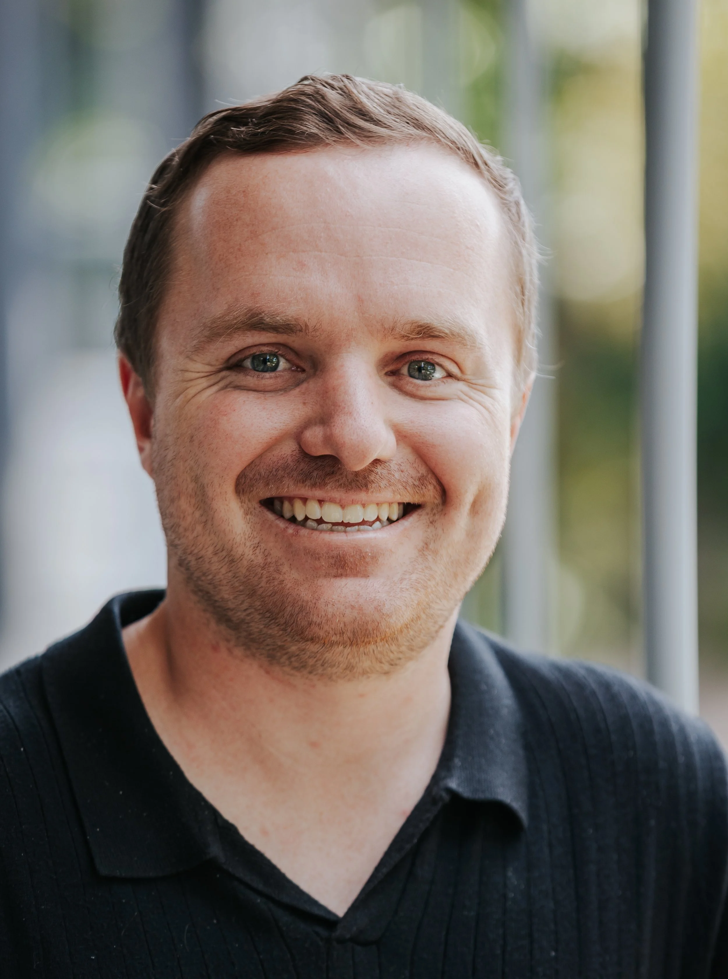 Close-up of a smiling man with short brown hair, blue eyes, and light skin, wearing a black sweater, with a blurred background.