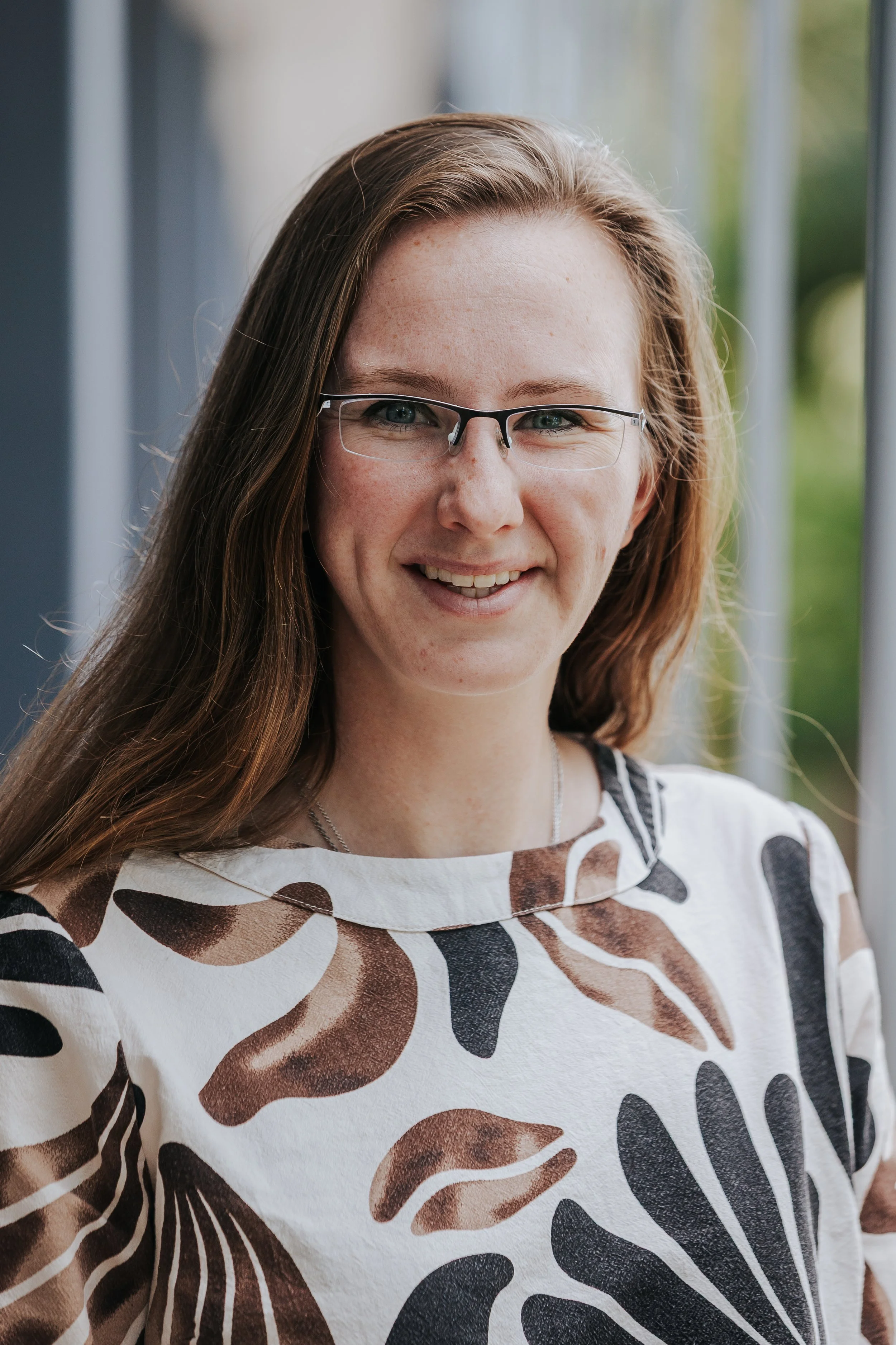 A woman with long brown hair, wearing glasses, smiling, standing near a window with green trees outside, wearing a white blouse with a large brown and black abstract pattern.