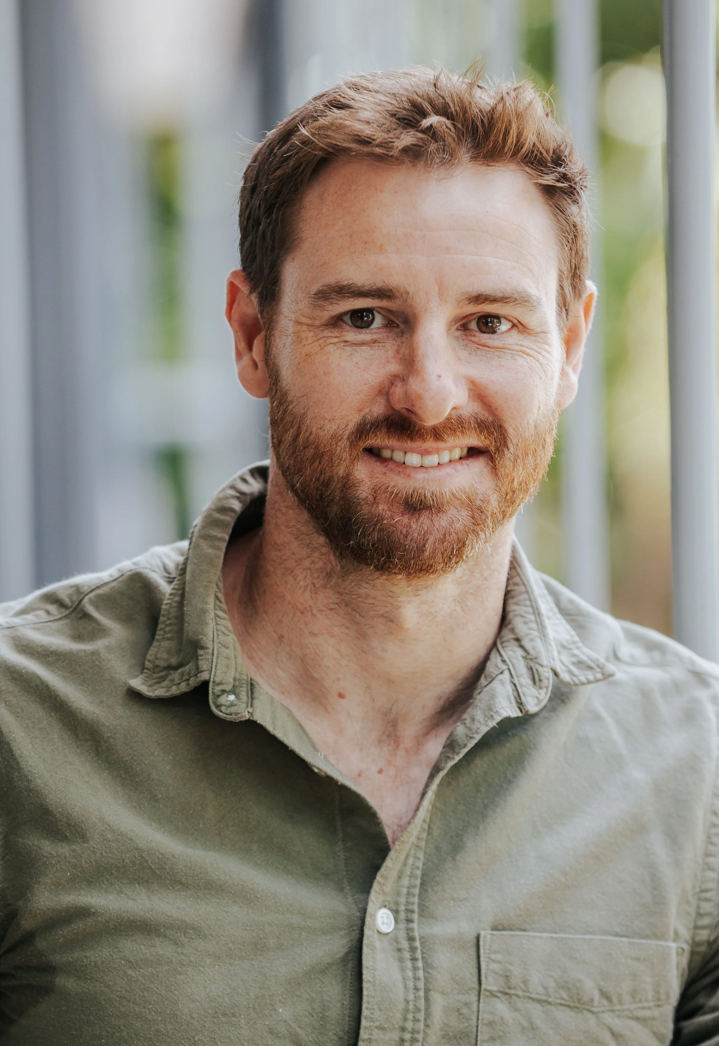 Close-up of a smiling man with light skin, red hair, and a beard, wearing a green button-up shirt with the top button open.