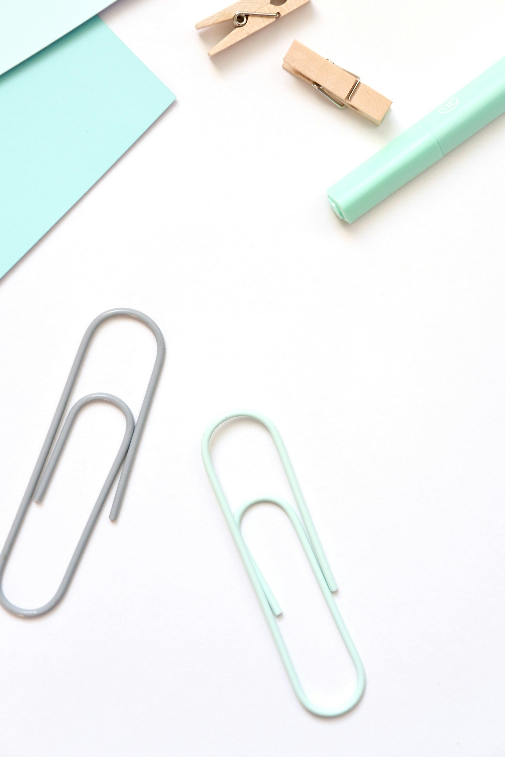 A background of office supplies including large paper clips, a small paper clip, a pastel blue sticker or notepad, a wooden clothespin, and a green dry erase marker on a white surface.
