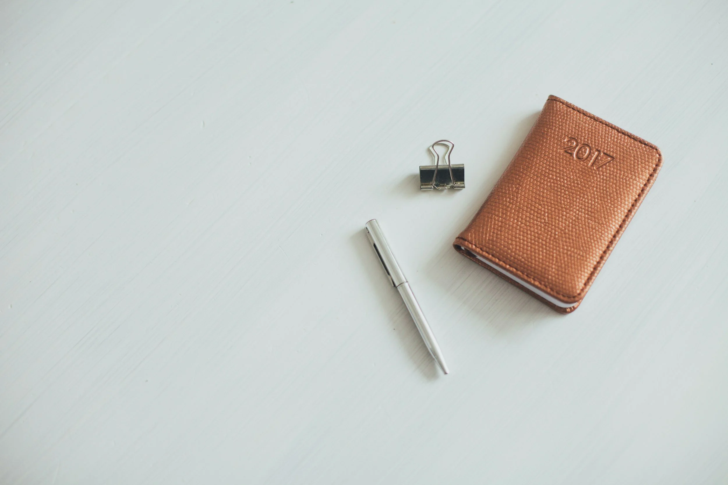 A white surface with a brown 2017 planner, a silver pen, a binder clip, and a metallic object.