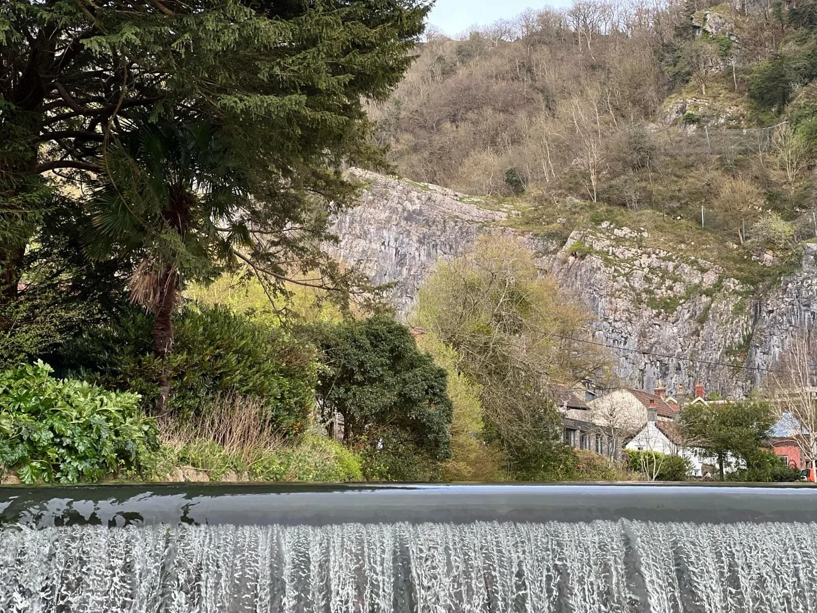 View of a waterfall at the bottom with greenery and trees on the sides, a rocky hillside in the background, and houses at the base of the hill.
