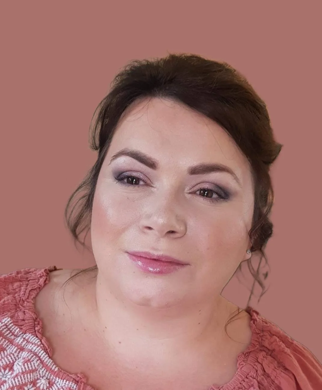 A woman with dark brown hair styled in loose curls, wearing makeup with pink lipstick, eyeshadow, and blush, and a pink top with white embroidery, smiling softly against a pink background.