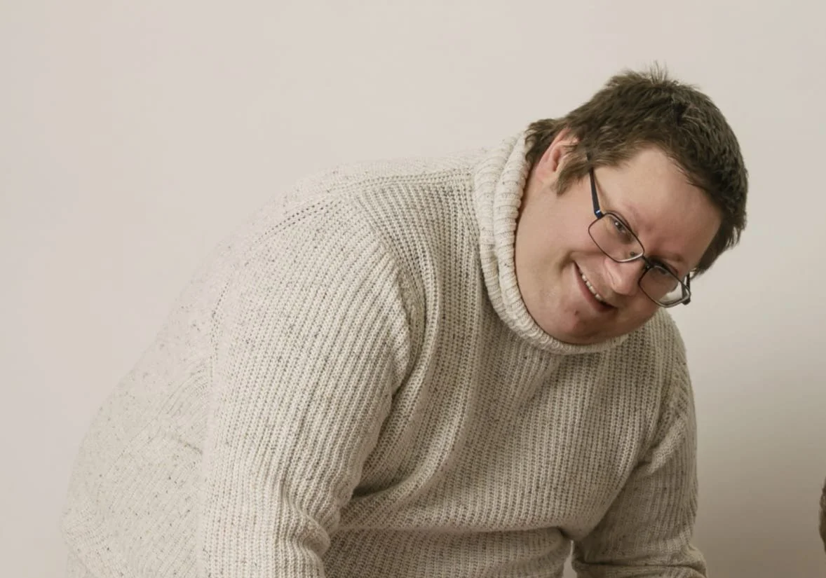 A man with glasses wearing a beige knit sweater, smiling and leaning forward.