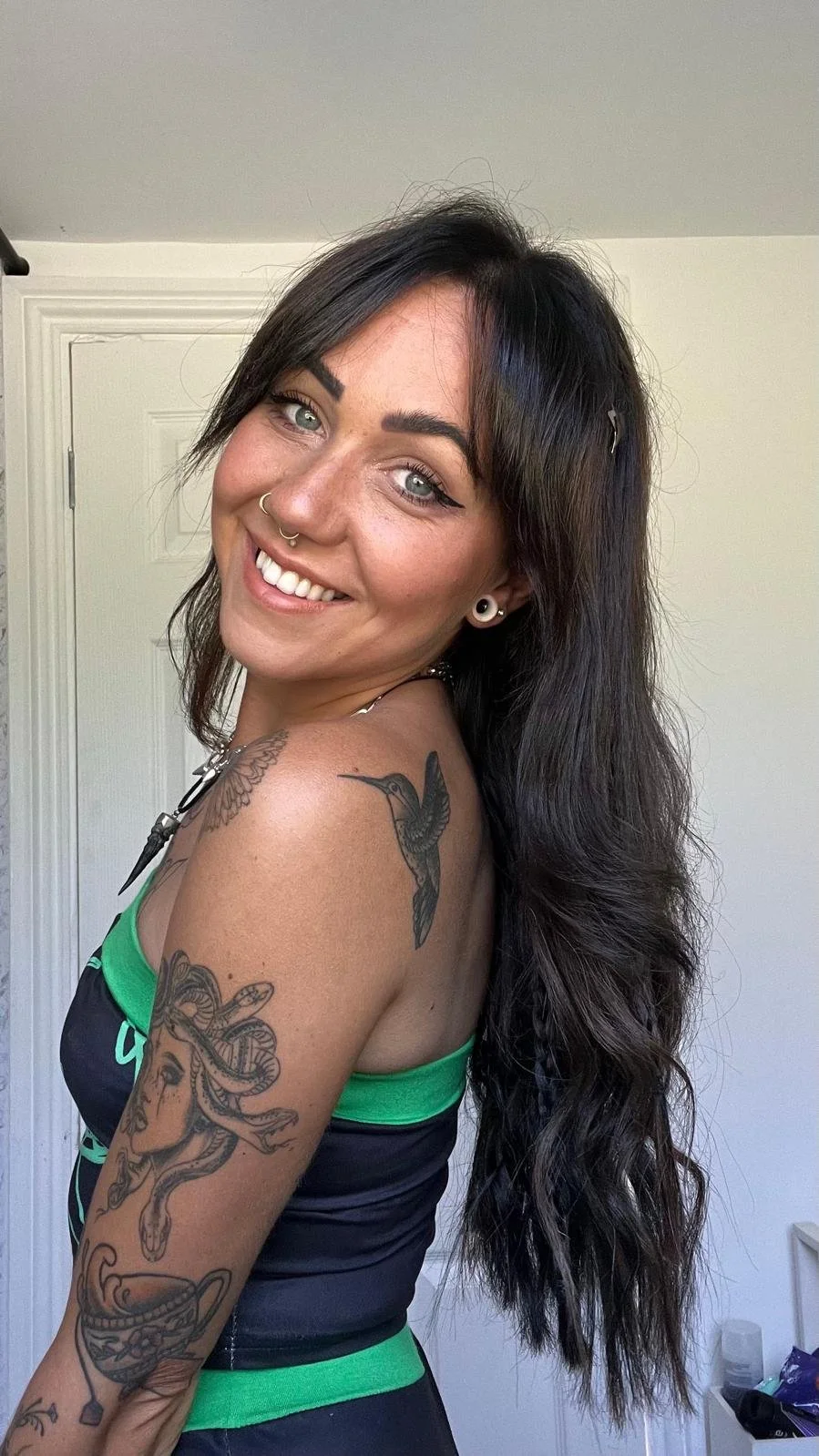 A woman with black hair, light skin, and tattoos, smiling and looking at the camera, standing in a room with a white door in the background.