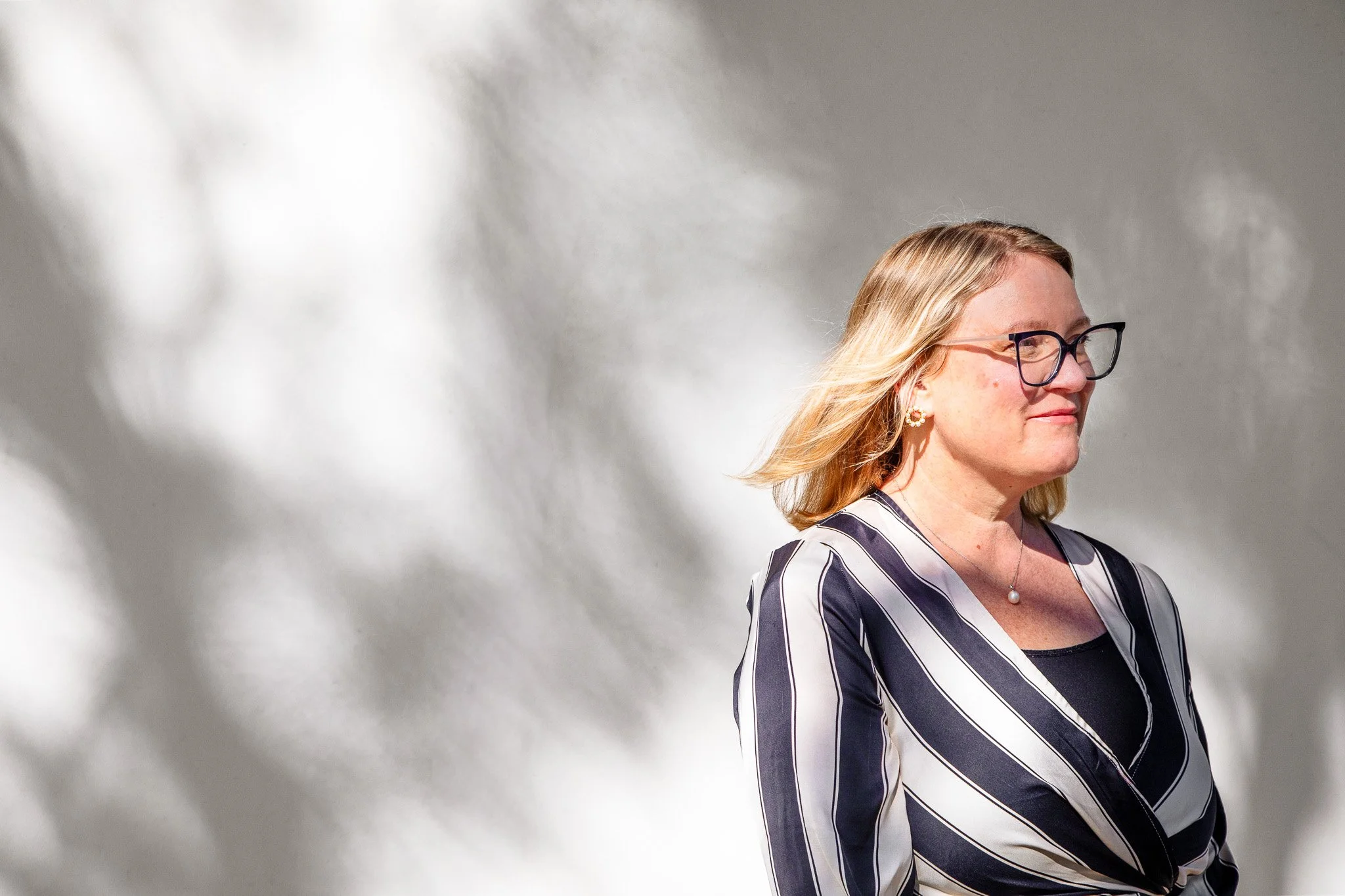 WordLab founder Hannah Edwards.  A woman with blonde hair, glasses, earrings, and a pearl necklace, wearing a black and white striped blazer, standing outdoors with sunlight and shadow pattern on her face and background.