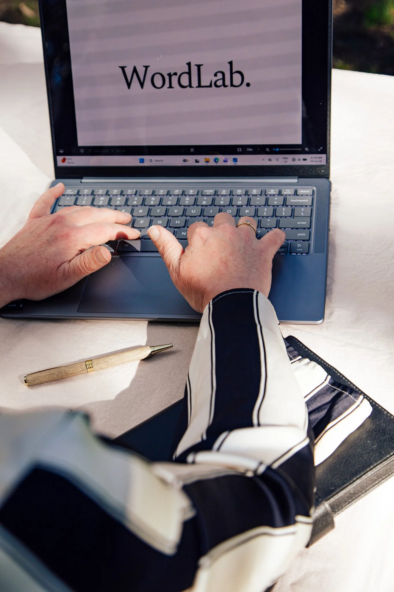 A person typing on a laptop keyboard, with a screen displaying 'WordLab.' The person is wearing a black and white striped sleeve, and the laptop is placed on a white surface with a gold-colored pen nearby.