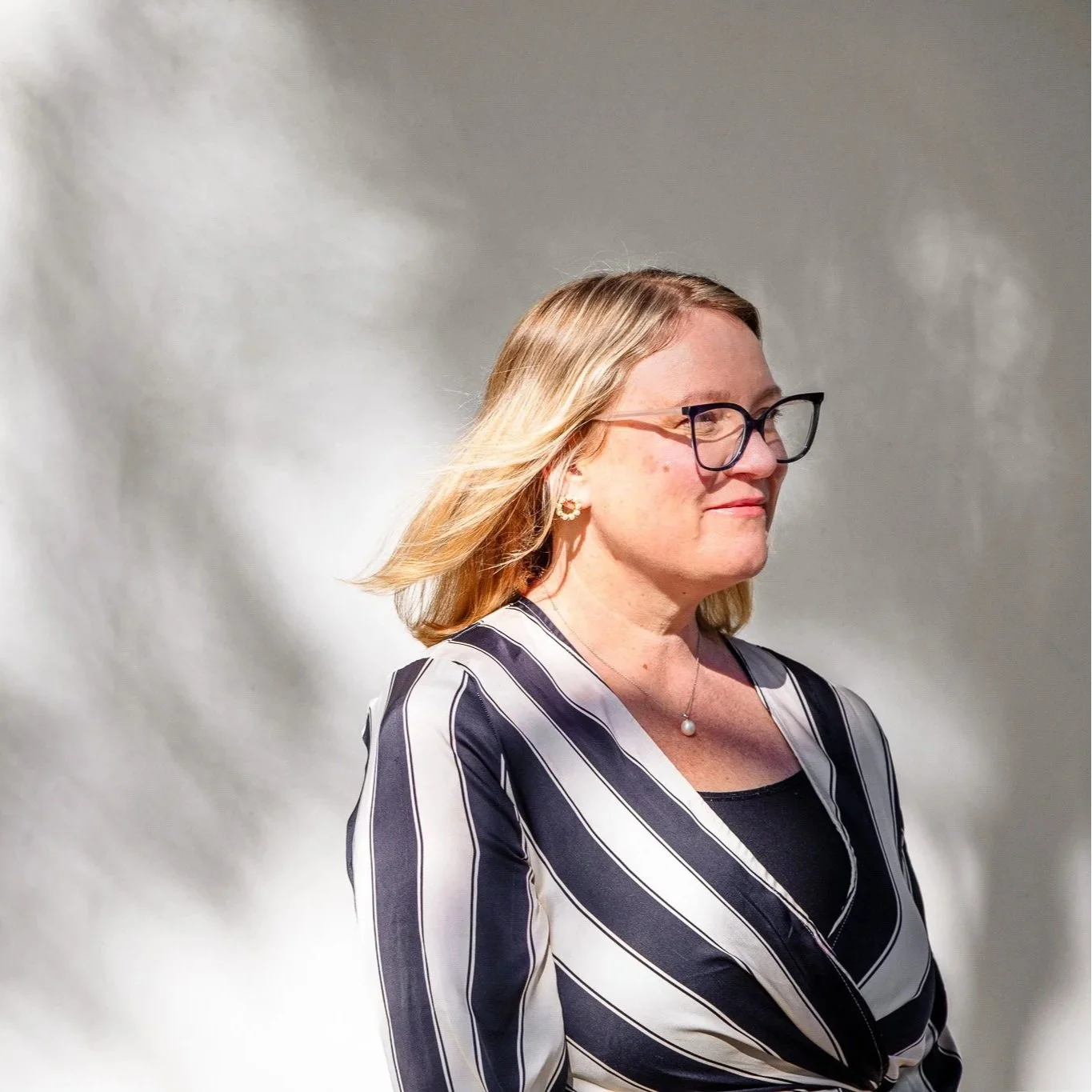 A woman with blonde hair, glasses, and earrings, wearing a striped blazer, standing sideways against a lightly textured background.