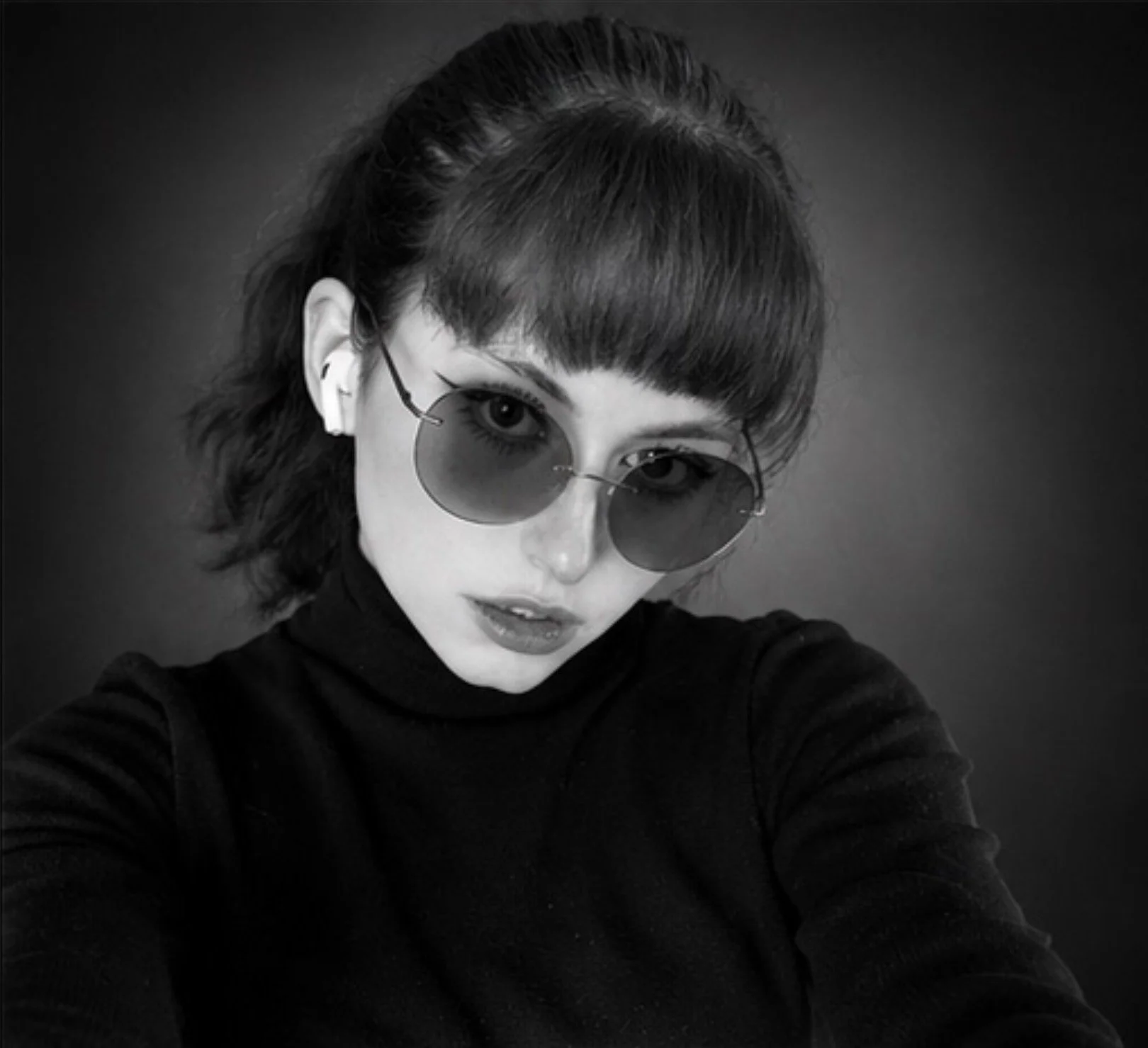 A woman with short dark hair, bangs, and glasses with round, tinted lenses, wearing a dark turtleneck, is taking a selfie against a plain dark background.
