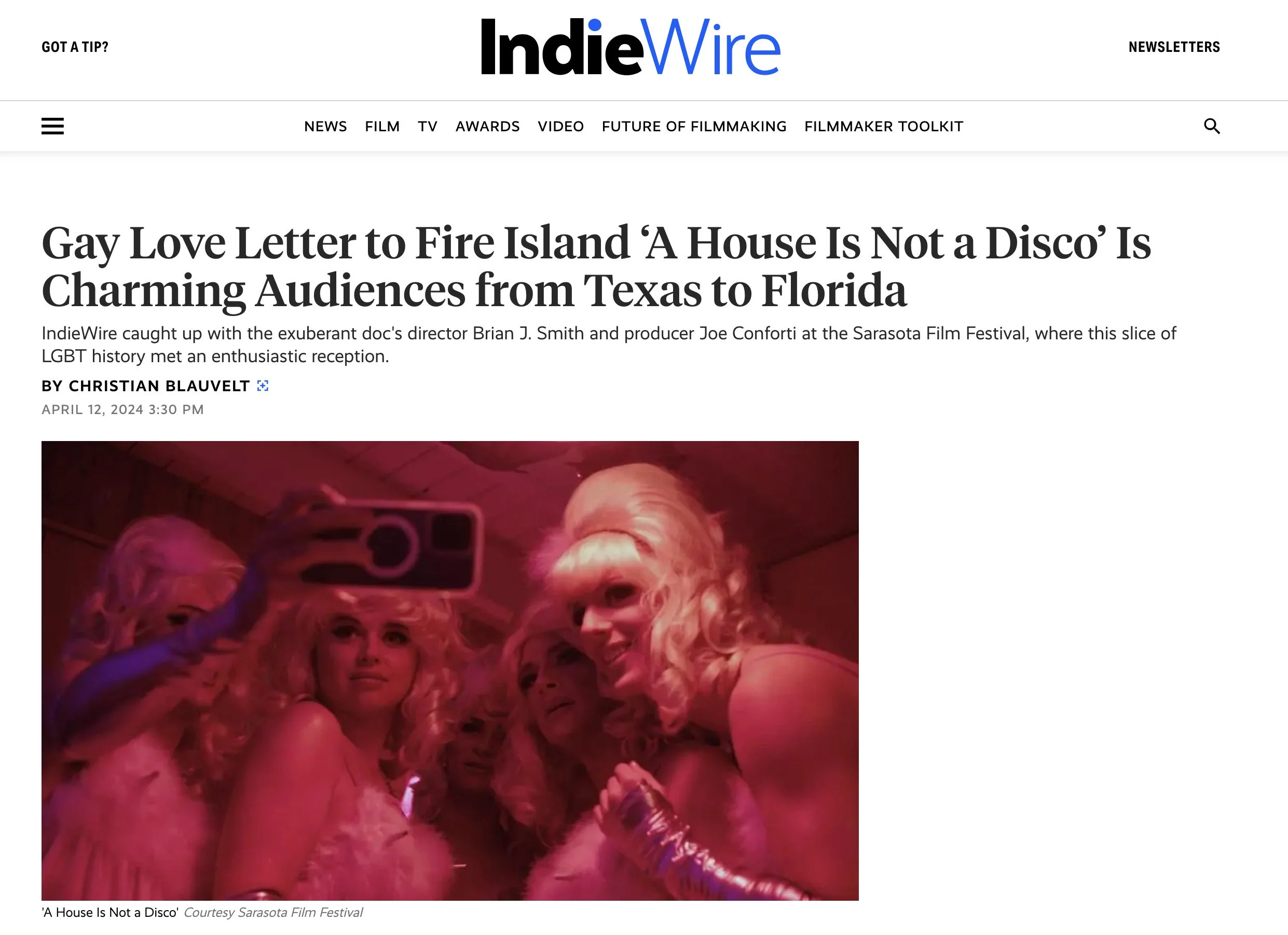 Indiewire Article about A House is Not a Disco - Picture: Group of gay men in drag and a trans women in elaborate costumes and makeup taking a selfie at a party, illuminated by pink lighting.