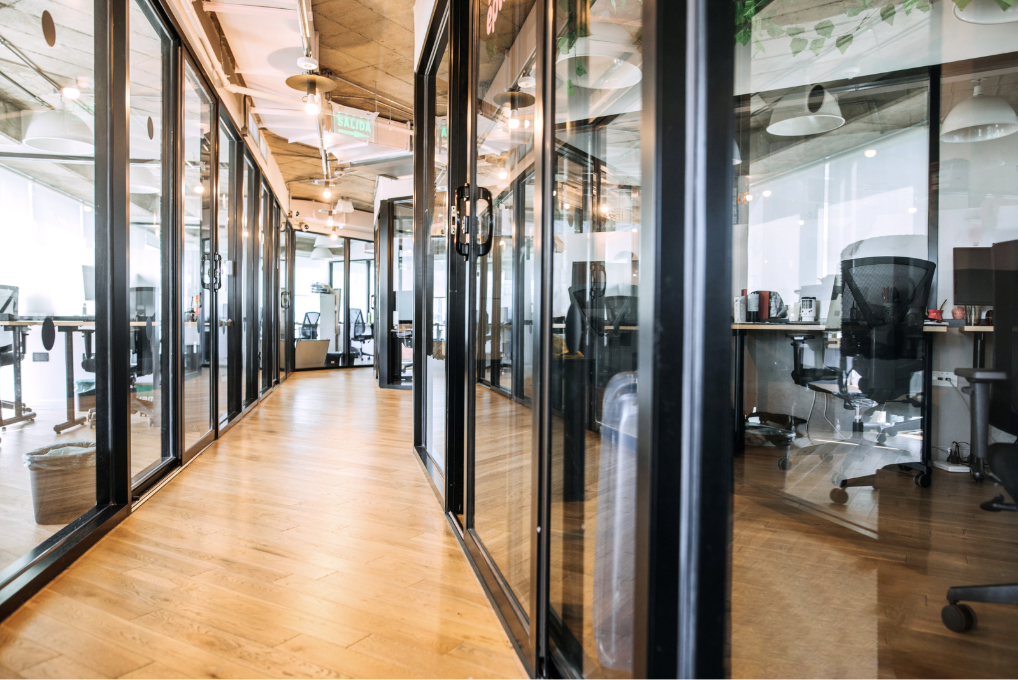 Modern office hallway with glass-walled private offices on both sides, wooden flooring, and gym equipment visible in the background.