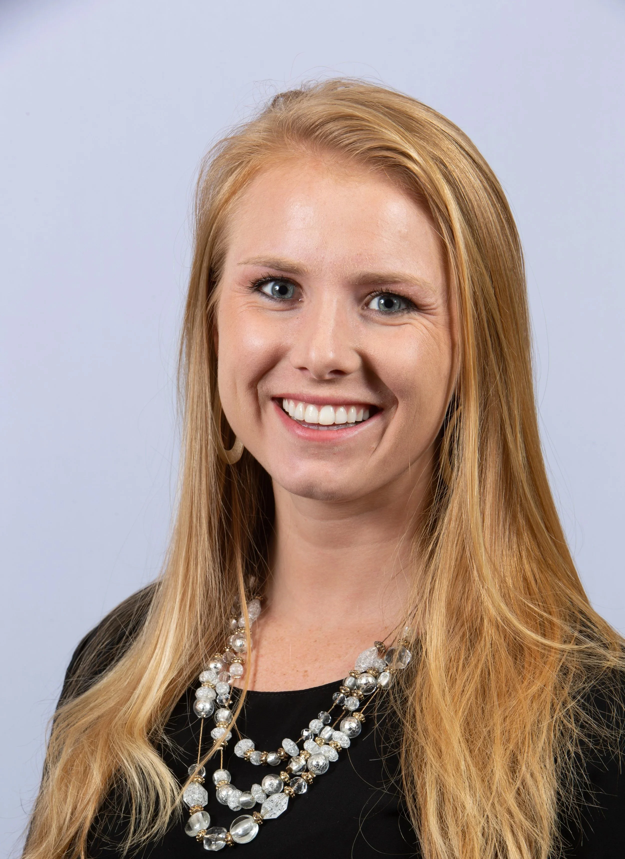 Close-up of a smiling woman with long blonde hair wearing multiple strands of large pearl and crystal necklaces, black top, and earrings, against a light background.