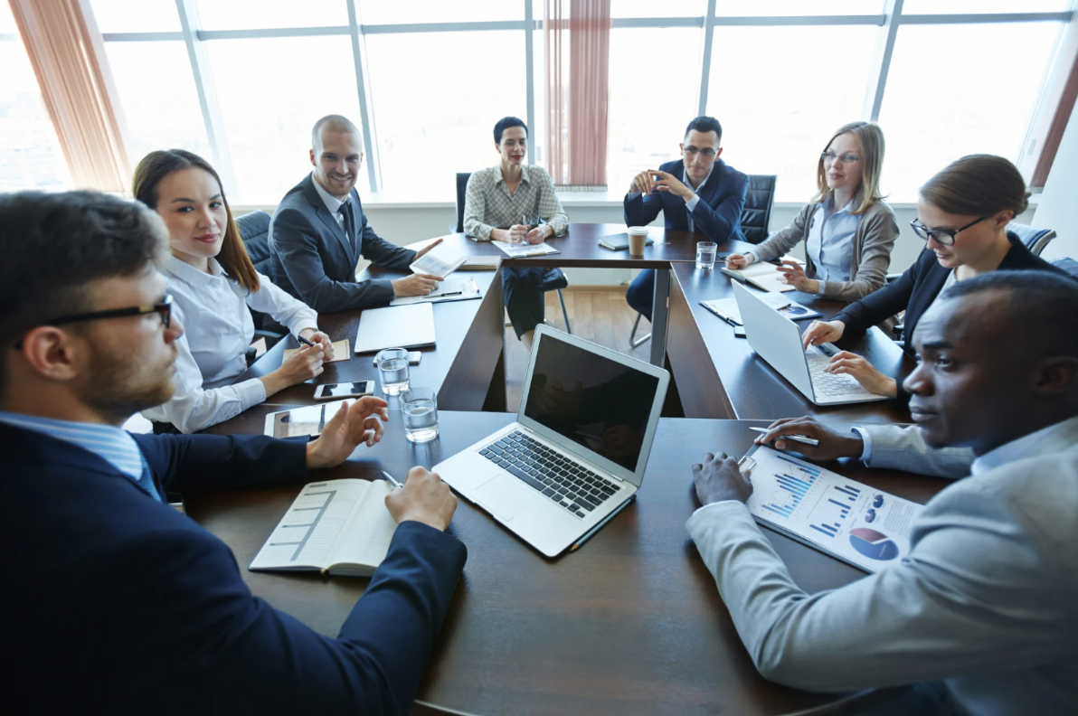 Business meeting with eight professionals sitting around a conference table in a modern office with large windows, some using laptops, others taking notes, and one person holding a smartphone.