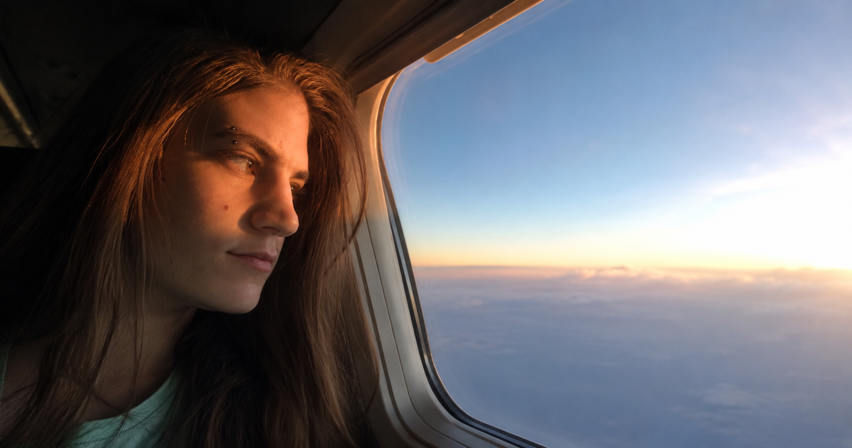 A young woman with long brown hair looking out the window of an airplane at the sky and clouds during sunset.