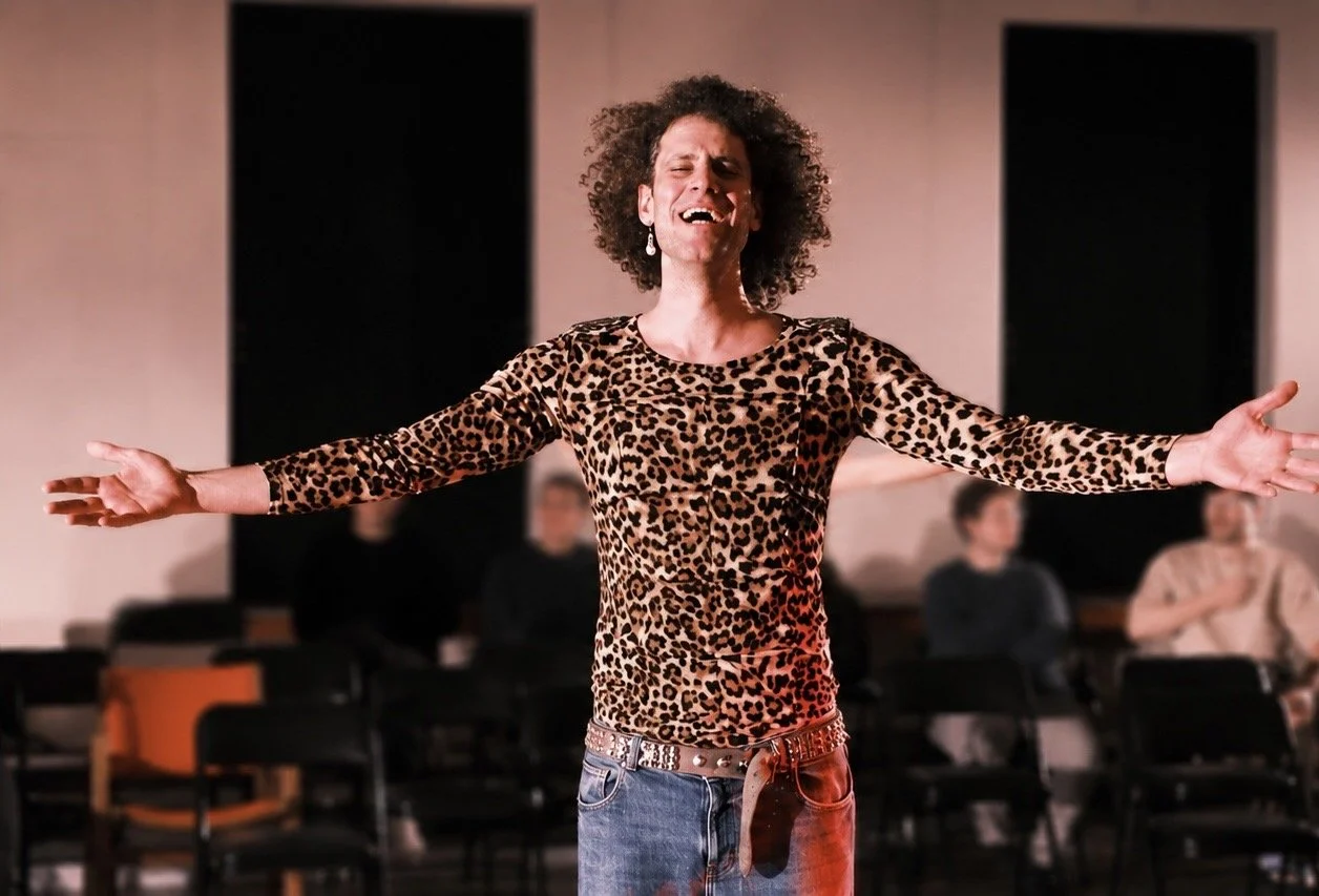 A woman with curly hair wearing a leopard print long sleeve shirt and jeans, singing with arms outstretched on stage, with a few seated audience members in the background.